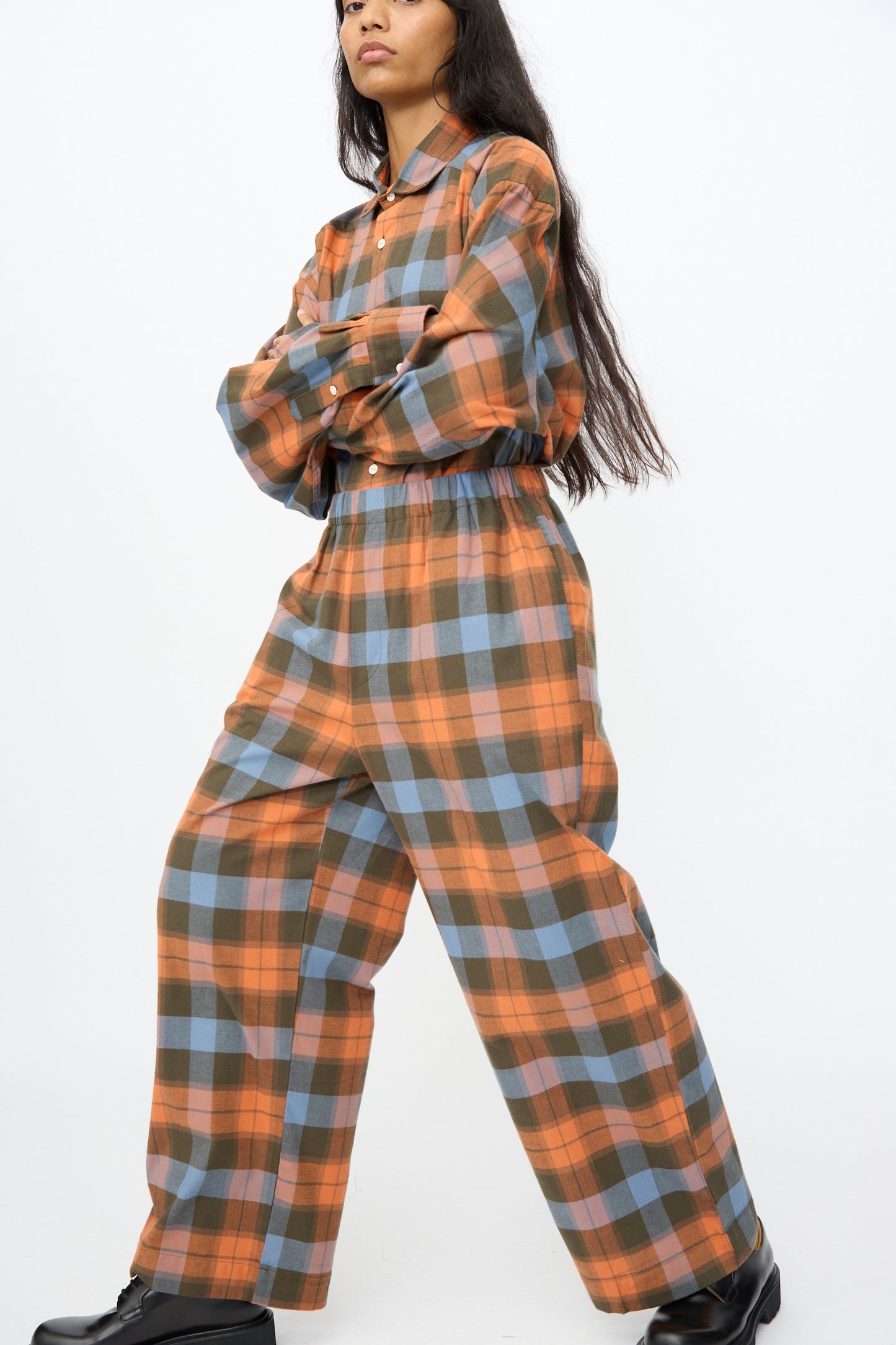 A person stands with arms crossed, wearing Baserange’s Claude Pant in Frame Plaid—loose-fitting, plaid wide leg pants—paired with a matching shirt in orange, blue, and gray tones and black shoes.