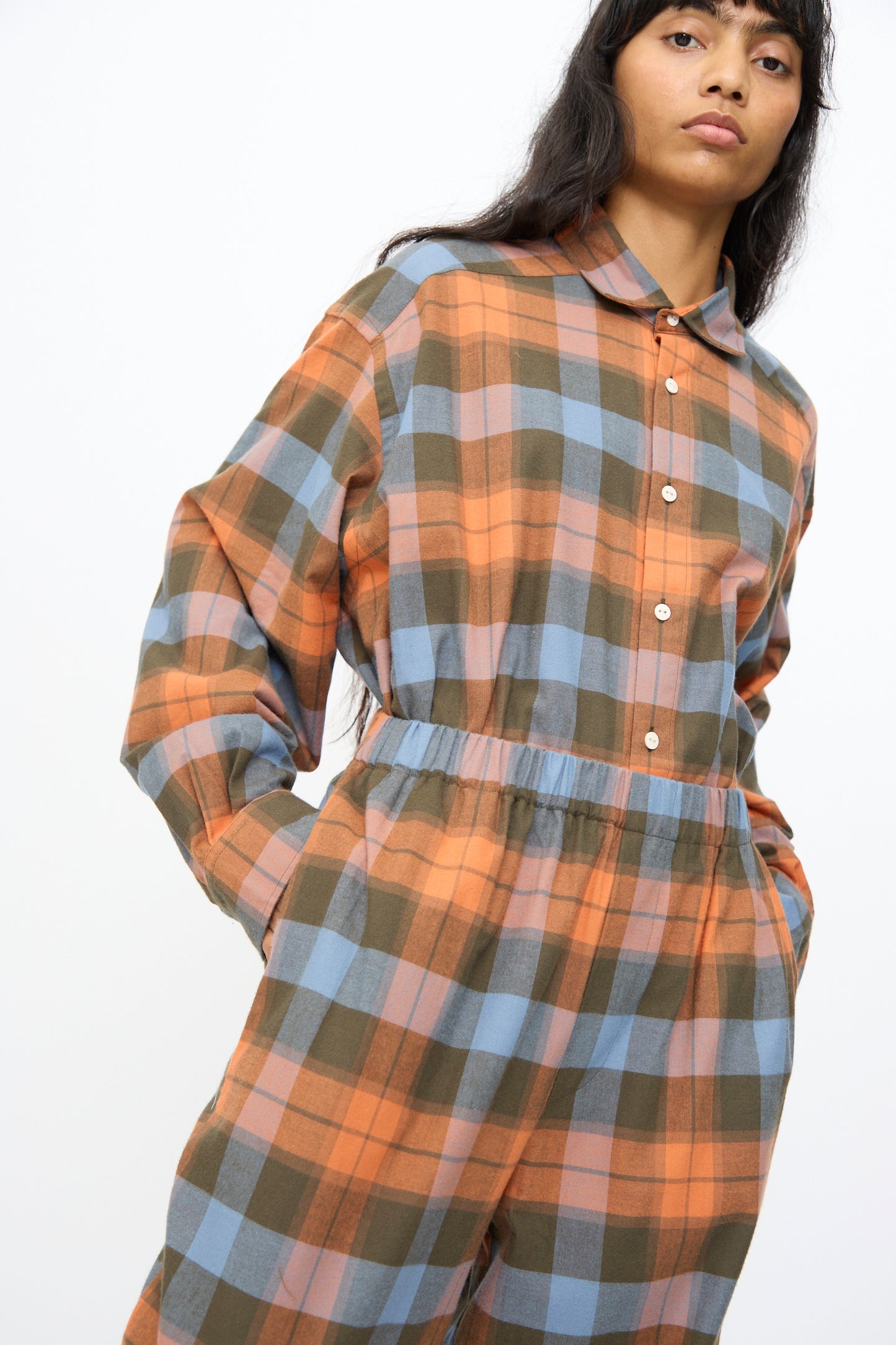 A person wears the Baserange Claude Pant in Frame Plaid—wide leg pants with an orange, blue, and olive green plaid pattern—standing against a plain white background.