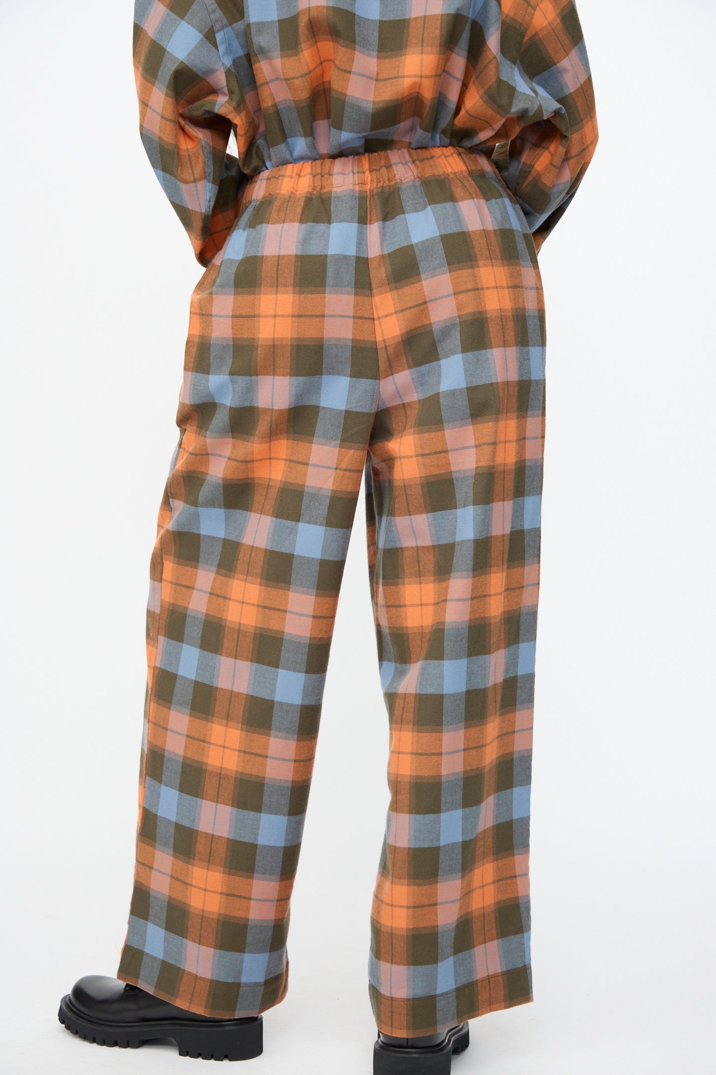 A person stands with their back to the camera wearing Baserange’s Claude Pant in Frame Plaid—wide leg pants featuring an orange, blue, and brown pattern—paired with visible black shoes.