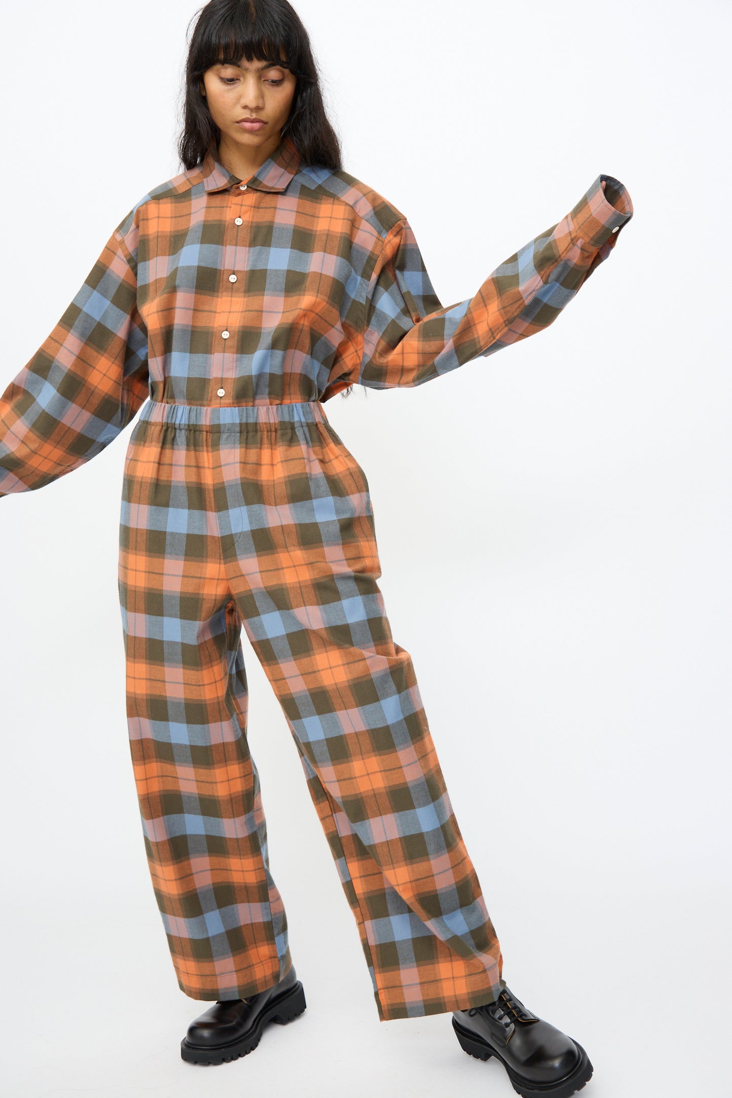 A model wears the Baserange Claude Pant in Frame Plaid, featuring a loose fit and wide legs in orange, blue, and brown plaid, styled with black platform shoes against a white background.