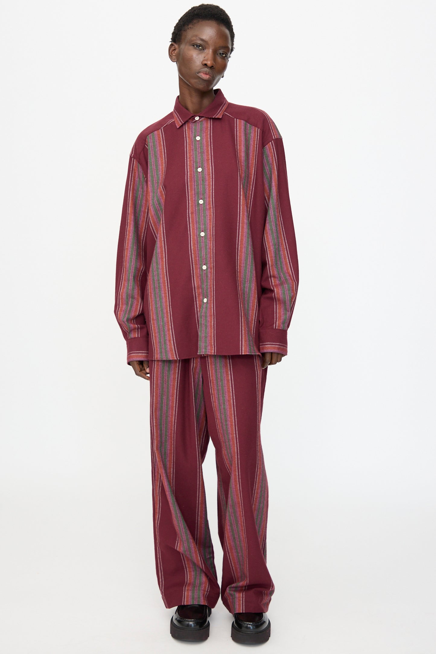 A person wears the Baserange Claude Shirt in Maroon Stripe, featuring long sleeves and a loose fit, paired with matching wide-leg pants, and stands against a plain white background.