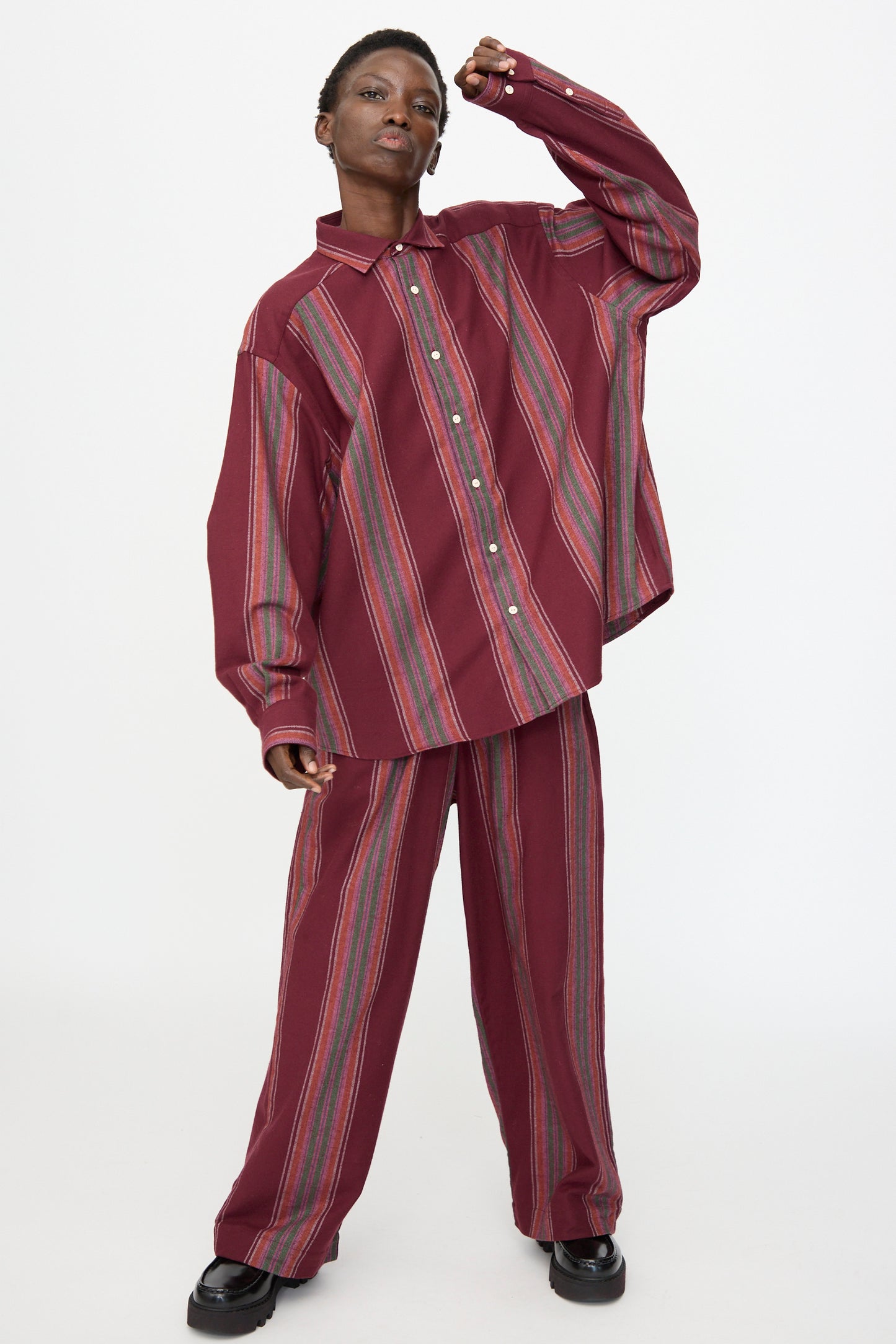 A person poses against a plain white background wearing the Baserange Claude Shirt in Maroon Stripe—an organic cotton, loose-fitting button-up—paired with matching wide-leg pants and black shoes.