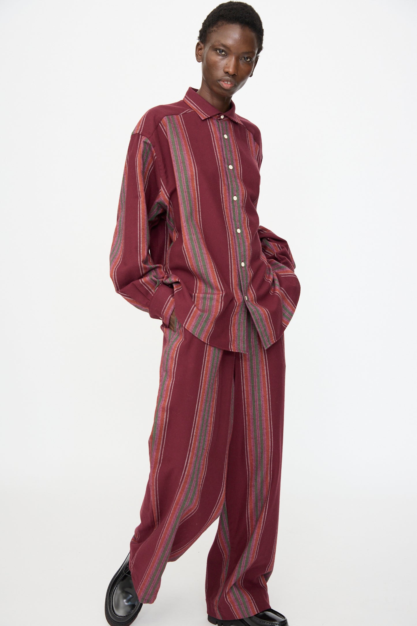 A person stands with hands in pockets against a white background, wearing Baserange's Claude Shirt in Maroon Stripe and matching wide-leg pants.