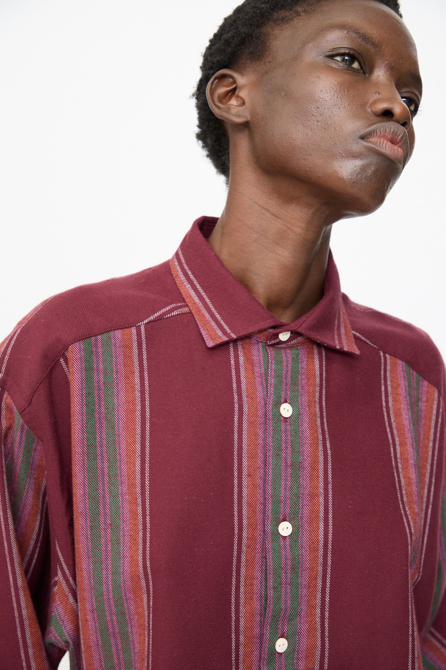 A person models the Baserange Claude Shirt in Maroon Stripe, featuring vertical colorful stripes, standing against a plain white background and looking slightly upward.