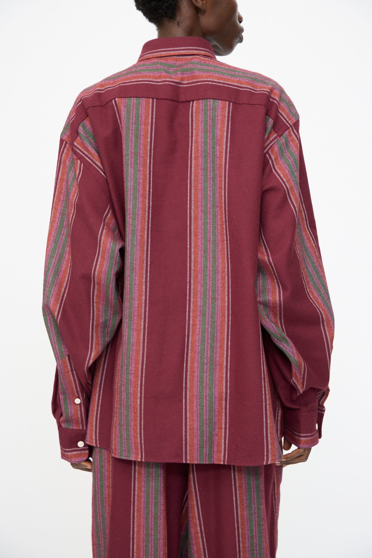 A person is shown from the back wearing the Baserange Claude Shirt in Maroon Stripe, a loose-fitting, long-sleeve organic cotton shirt with multicolored vertical accents; matching pants are partially visible.