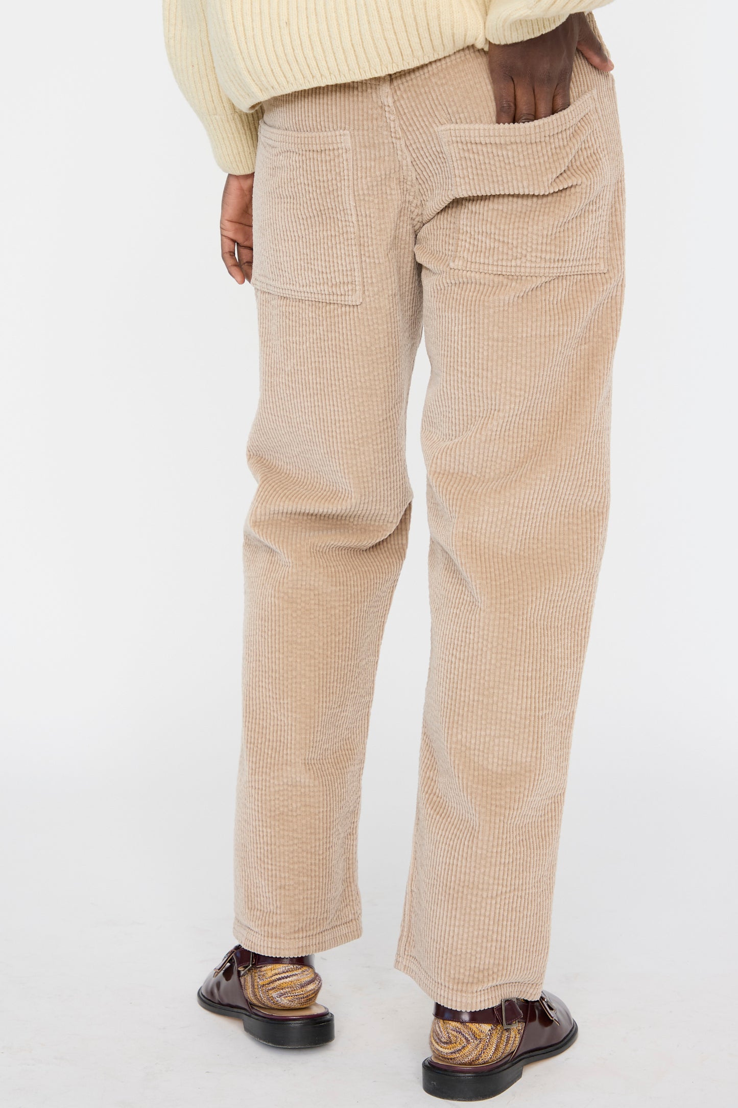A person wearing Baserange Corduroy Indre Pant in Sand and brown shoes is seen from the back with a hand in the pocket against a plain backdrop.