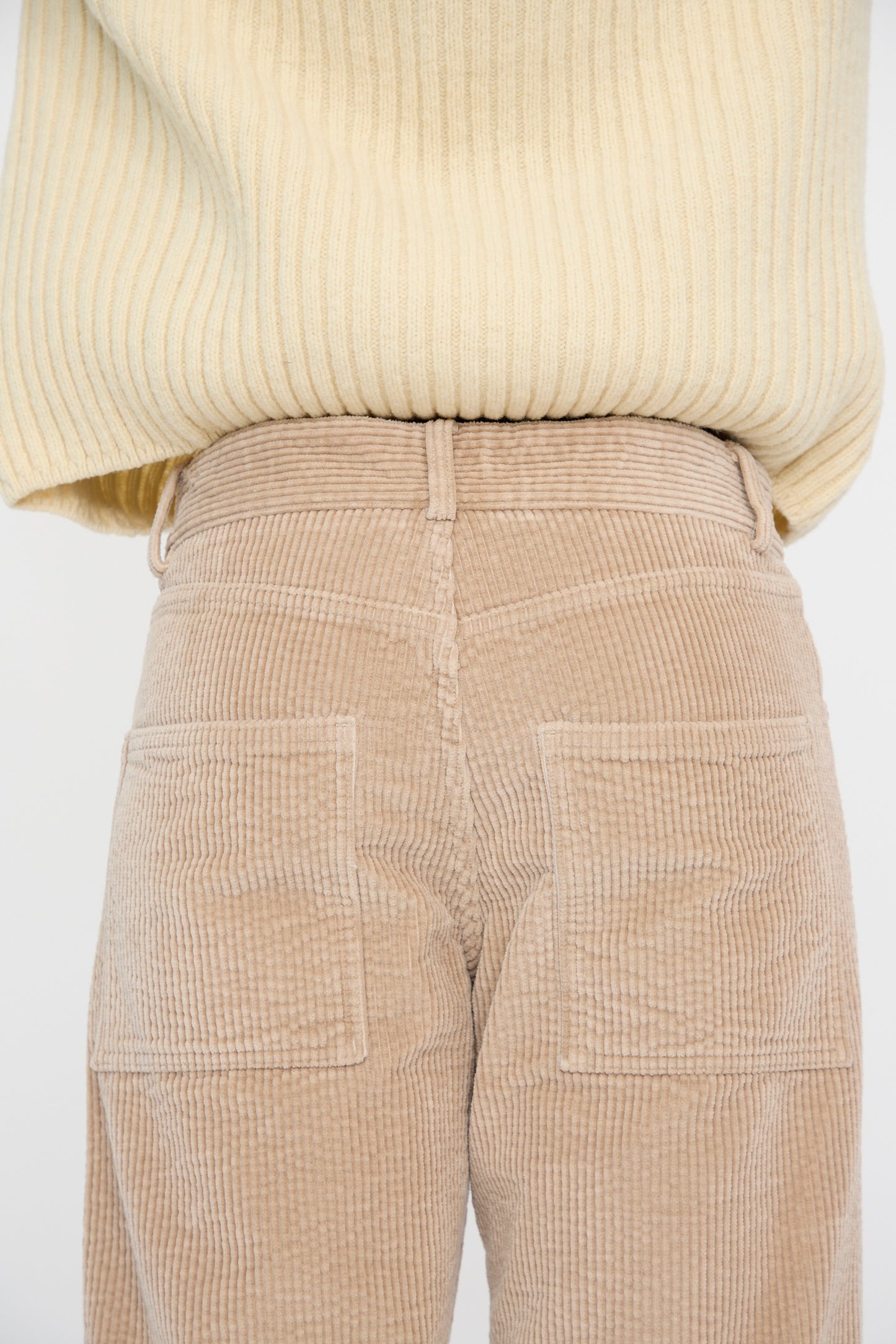 A person is facing away wearing a vertically ribbed beige sweater and Baserange Corduroy Indre Pant in Sand, featuring visible back pockets, closeup on the hips.
