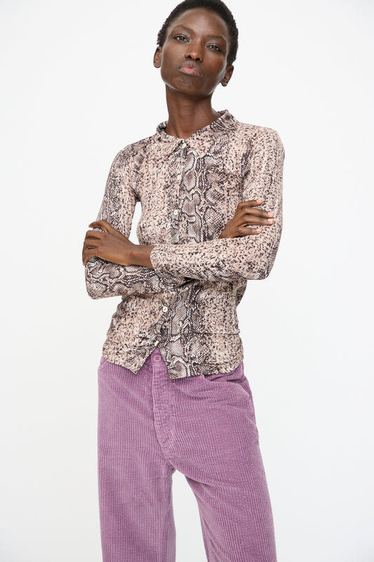 A person stands with arms crossed, wearing the Baserange Cosme Shirt in Snake—a slim fit, snakeskin-patterned long sleeve—paired with lavender corduroy pants against a plain white background.