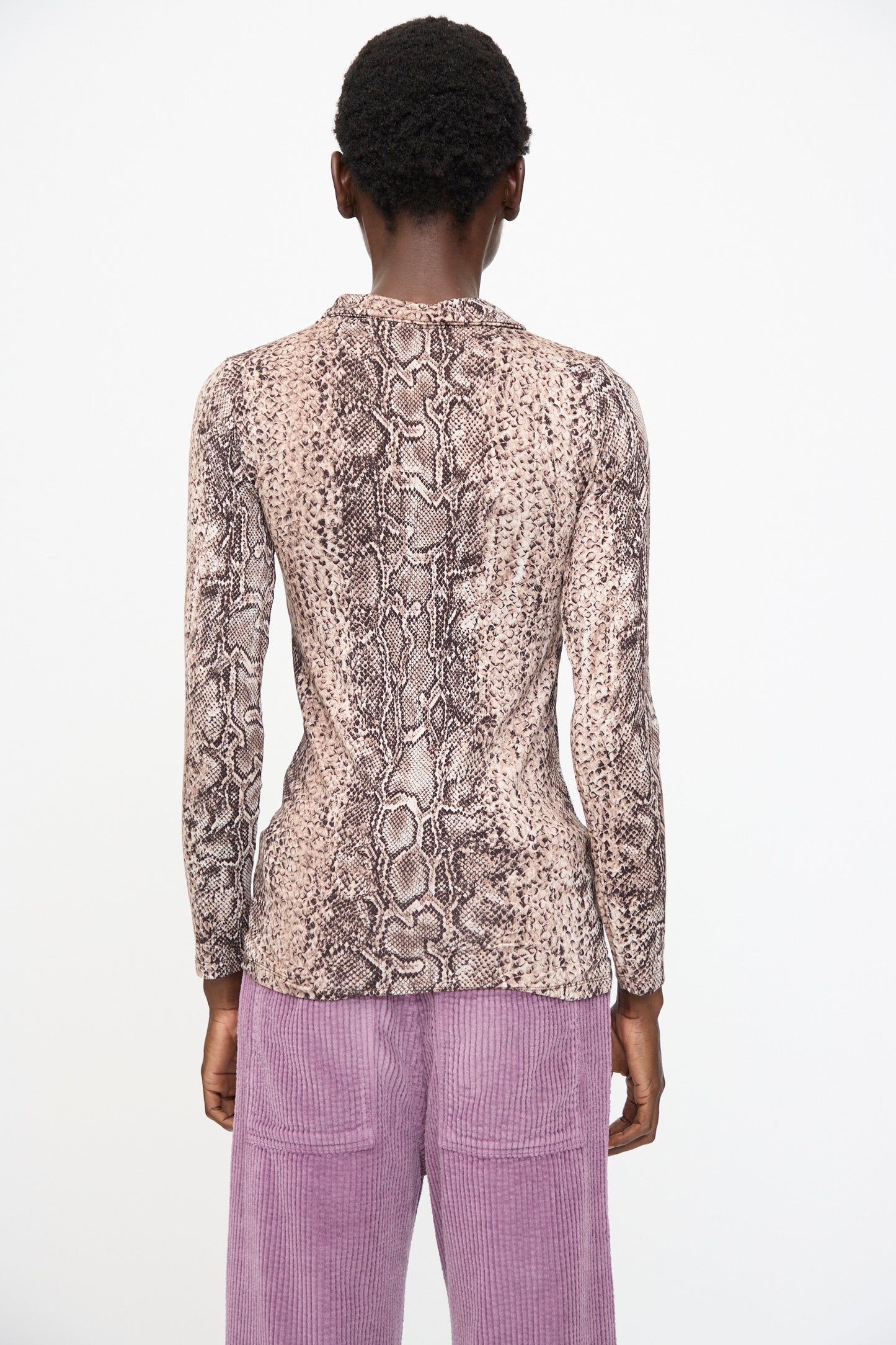 A person faces away, wearing the Baserange Cosme Shirt in Snake, a slim fit brown snakeskin-patterned long sleeve shirt, paired with light purple corduroy pants against a plain white background.