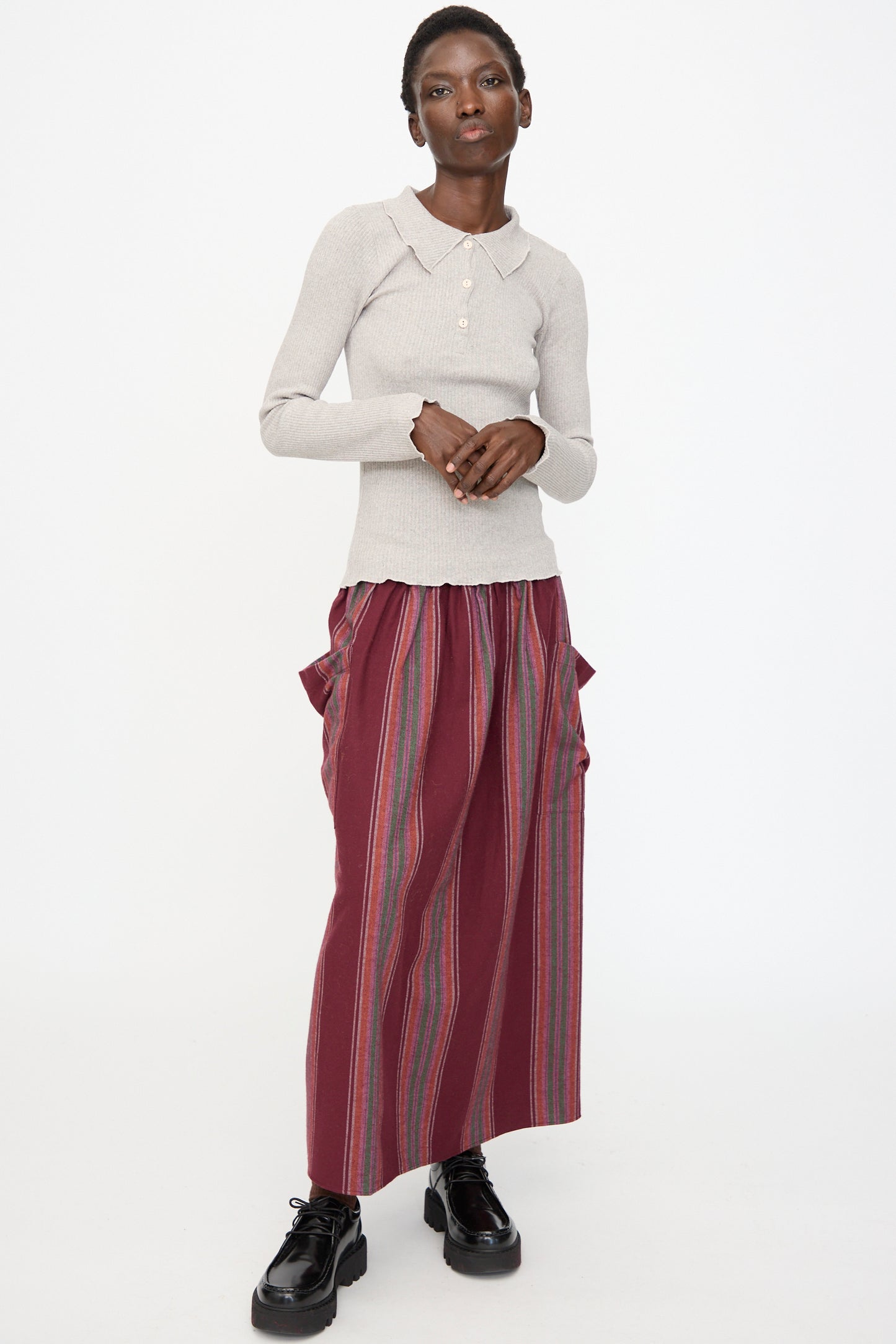 A person stands against a white background wearing the Baserange Dual Long Sleeve Tee in Grey Melange with a long maroon skirt featuring vertical stripes and black shoes.