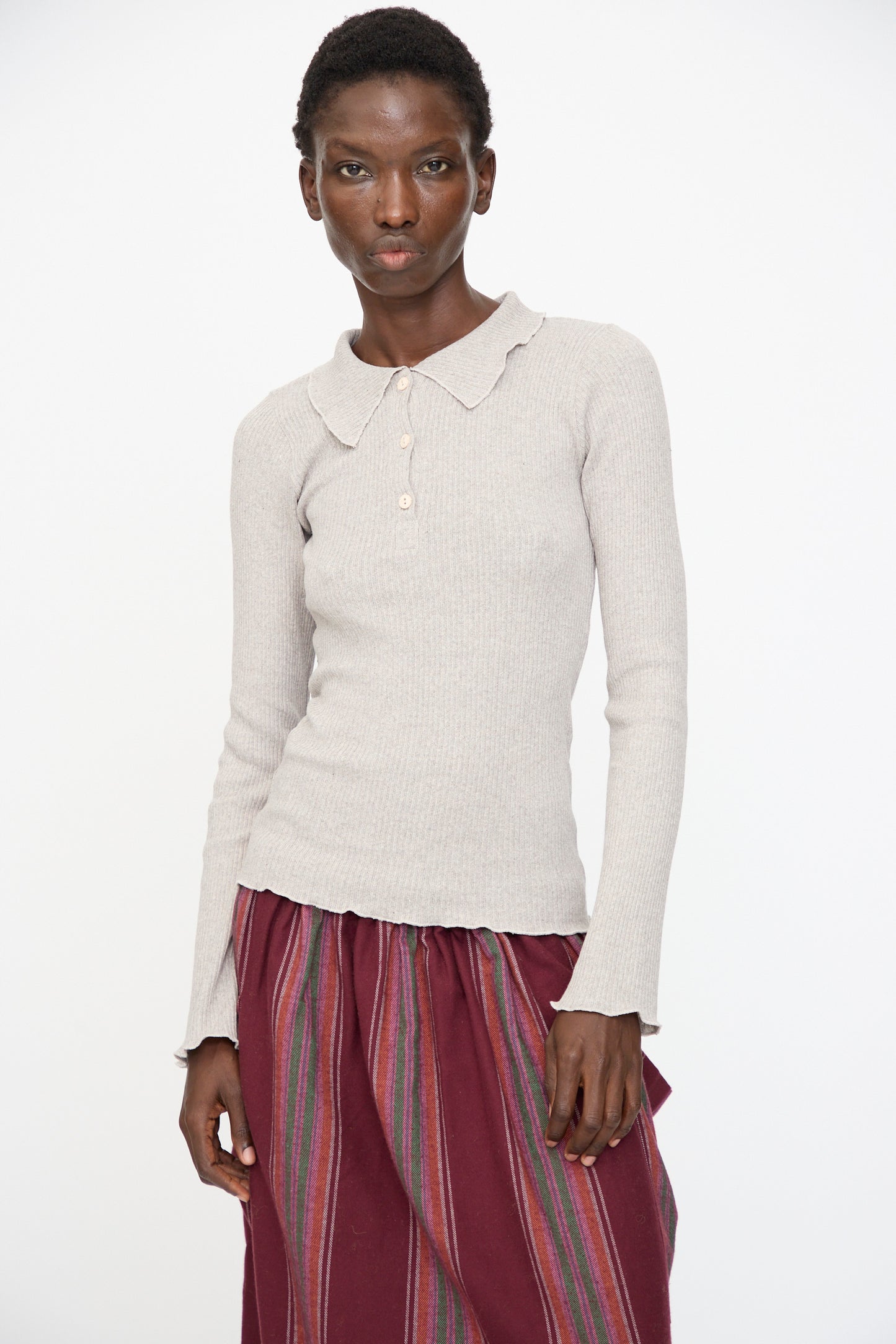 A person stands against a plain background wearing the Baserange Dual Long Sleeve Tee in Grey Melange, paired with a maroon skirt featuring vertical multicolored stripes.