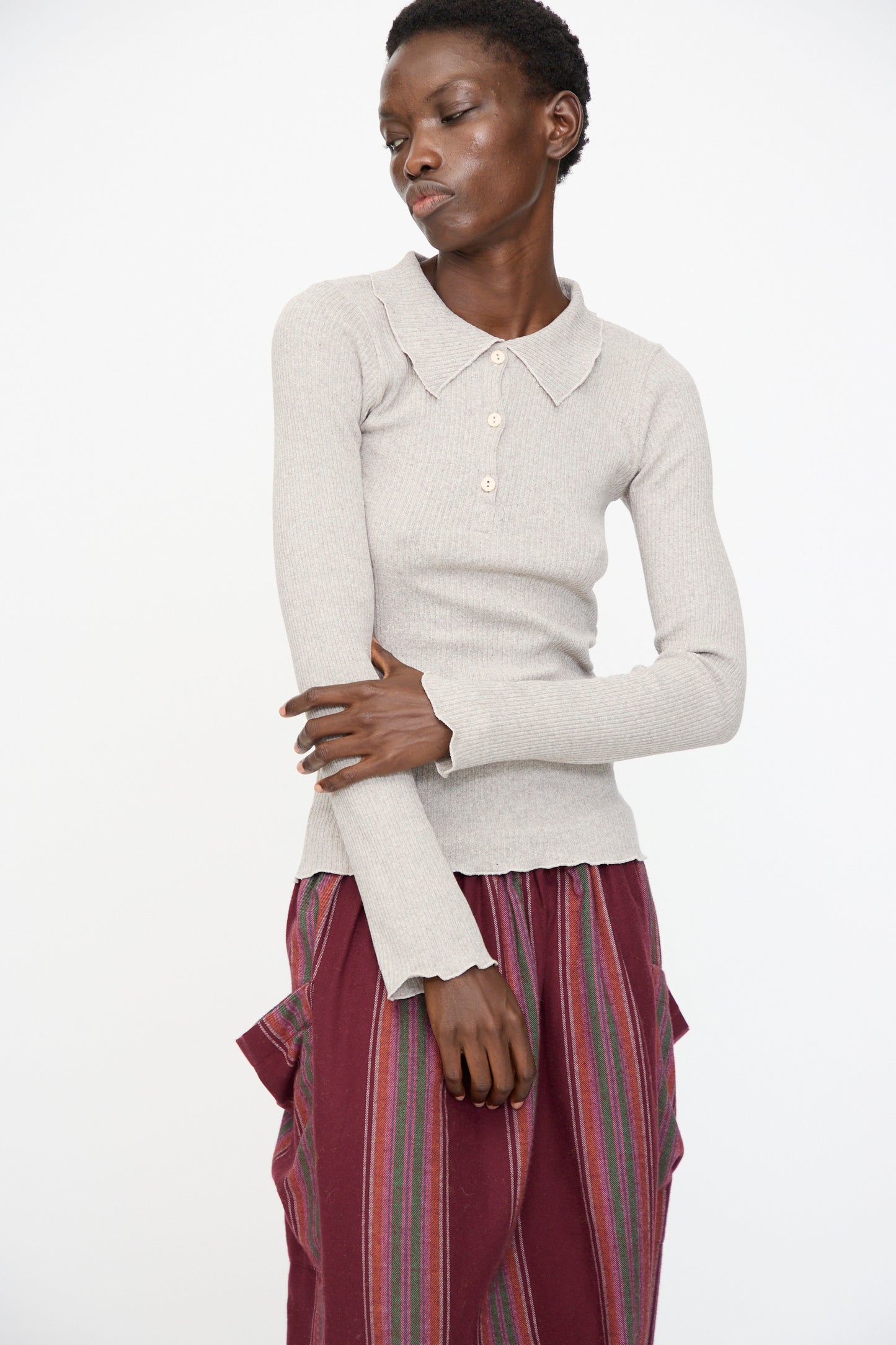A person wears the Baserange Dual Long Sleeve Tee in Grey Melange, styled with a burgundy striped skirt with side pockets, standing against a plain white background.