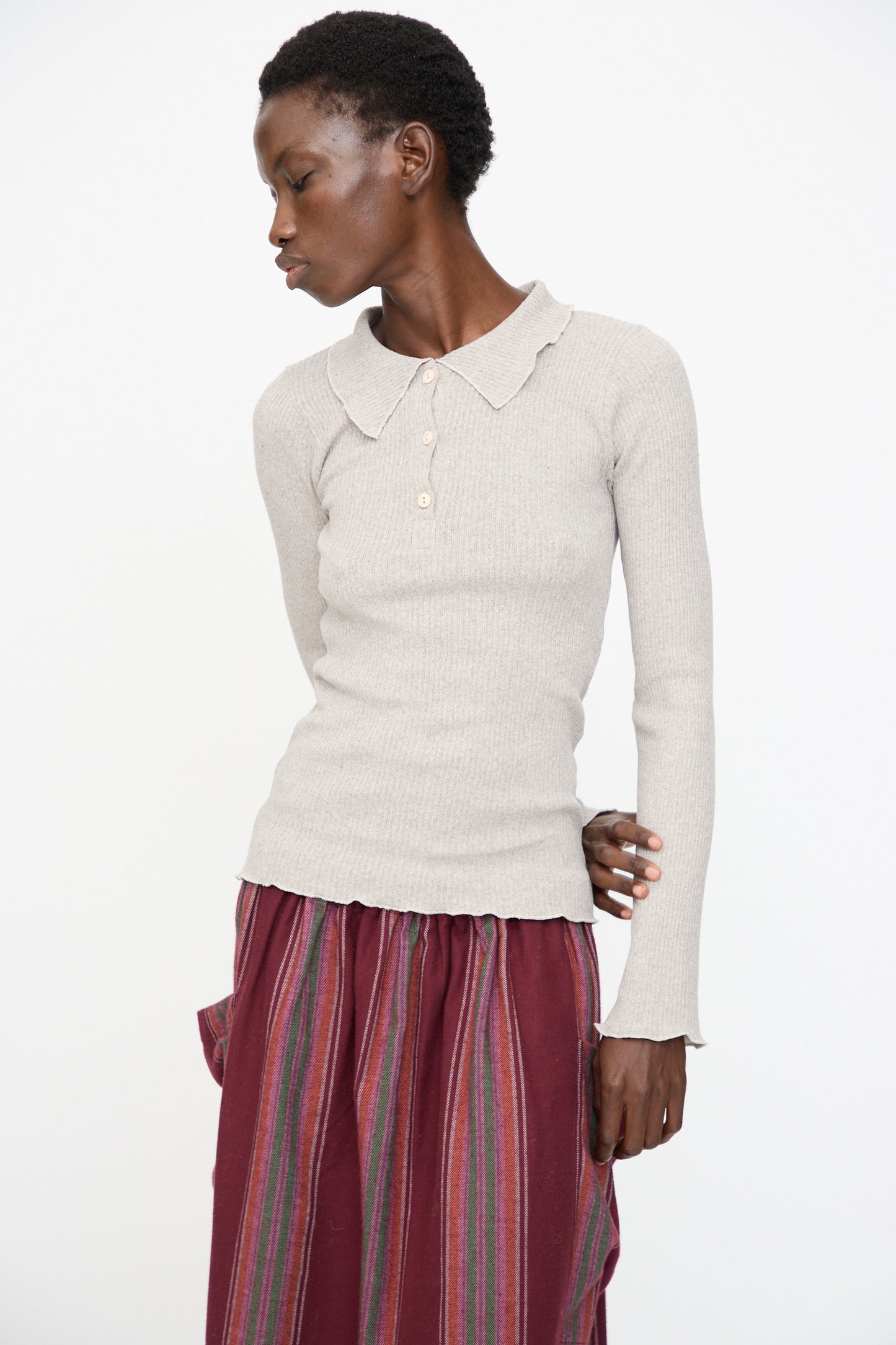 A person wearing the Baserange Dual Long Sleeve Tee in Grey Melange paired with a maroon skirt with multicolored vertical stripes stands against a plain white background.