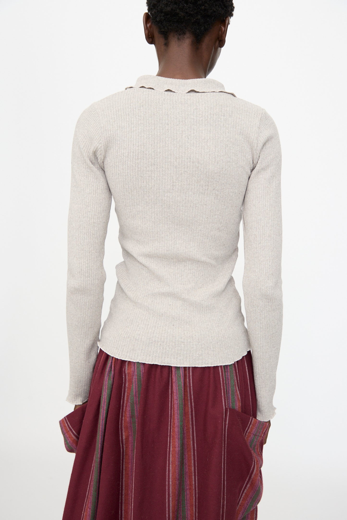 Person stands with their back to the camera, wearing the Baserange Dual Long Sleeve Tee in Grey Melange, a slim fit top made from recycled cotton, paired with a maroon skirt featuring multicolored vertical stripes.