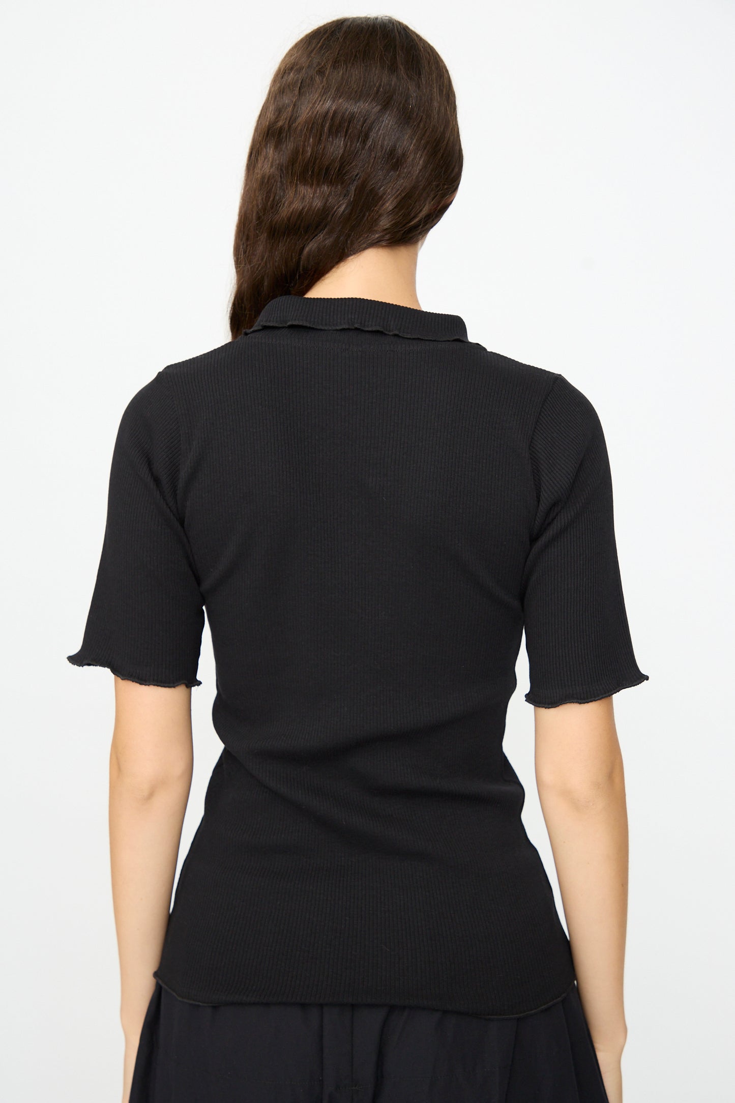 A woman with long brown hair is photographed from the back against a plain white background, wearing the Baserange Dual Polo in Black—a ribbed, slim fit short-sleeve shirt.