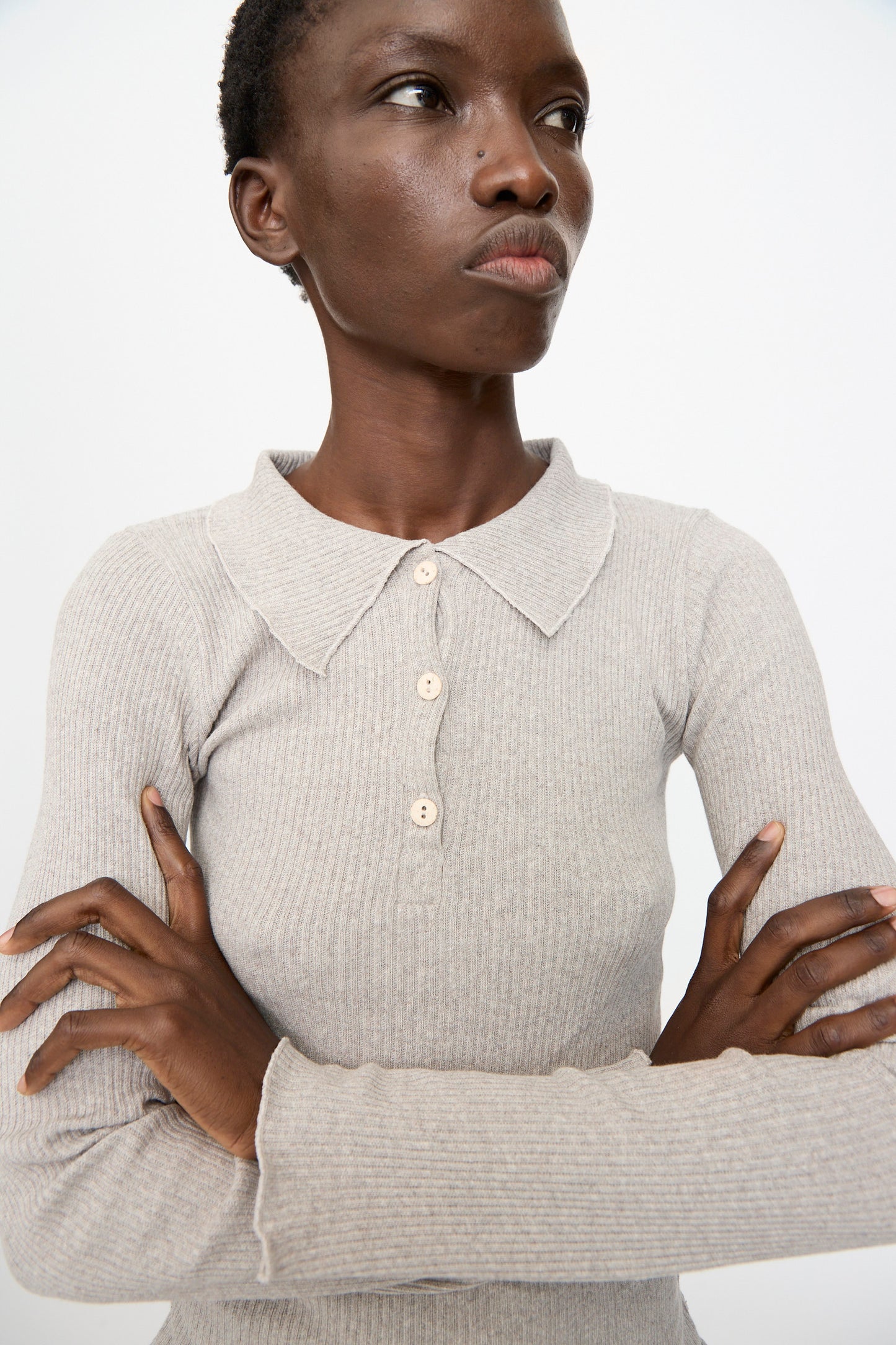 Someone stands with arms crossed against a plain background, wearing the Baserange Dual Long Sleeve Tee in Grey Melange—a slim fit, ribbed shirt featuring a collar and button details.
