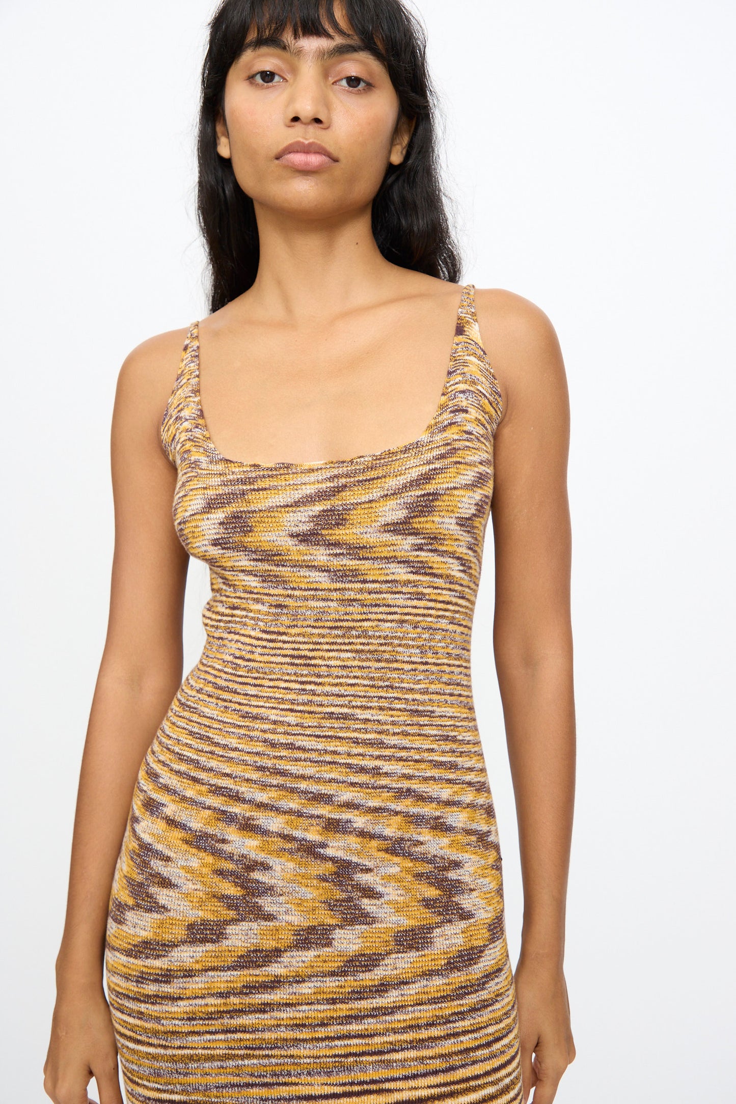 A woman with long dark hair wears the Baserange Flute Dress in Brown Melange, a slim-fit knit midi dress with a scooped neckline, standing against a plain white background.
