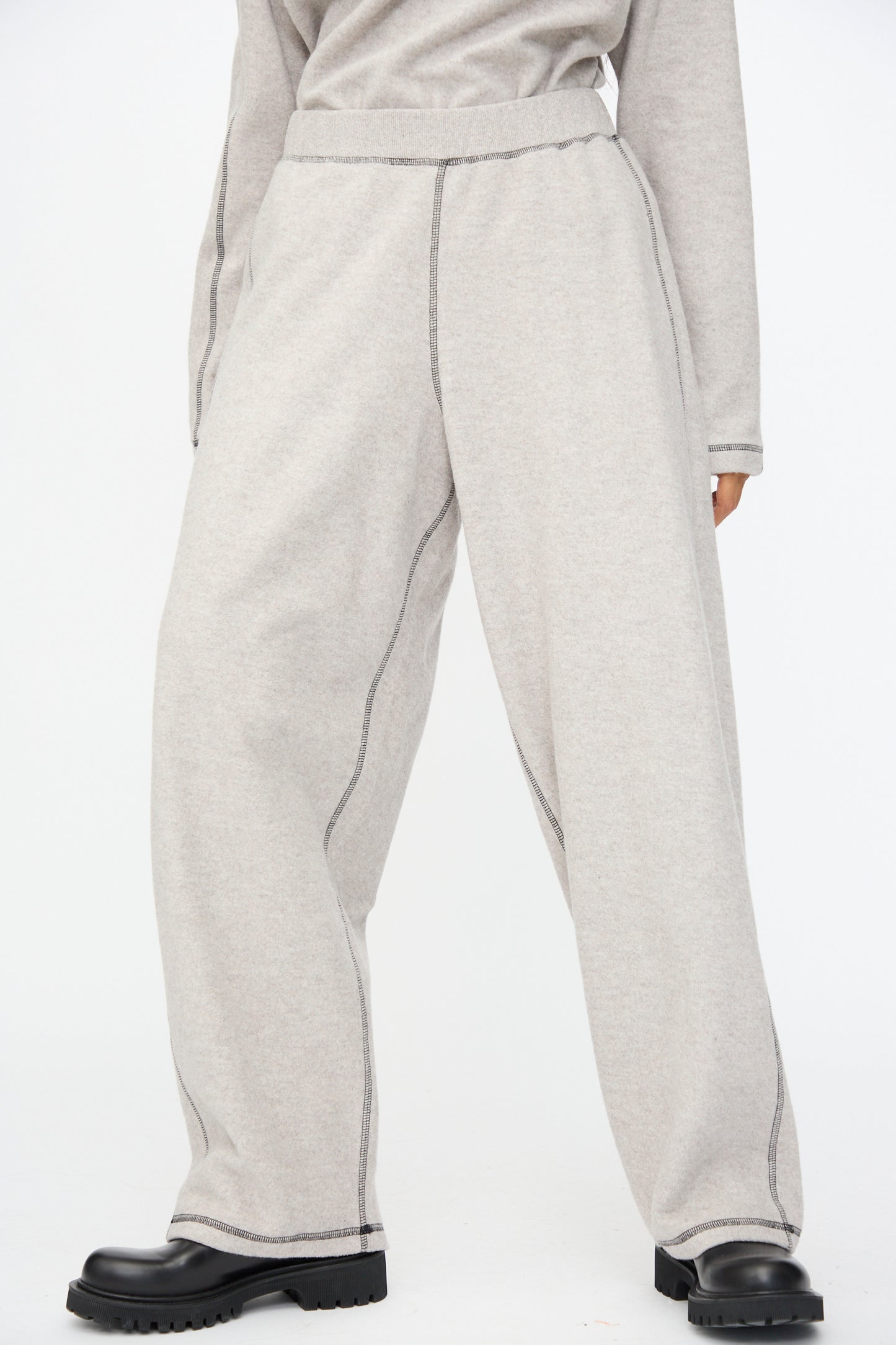 Person wears Baserange Fold Sweatpant in Grey Melange—wide-leg, recycled cotton pants with visible dark seams—paired with a matching top and black chunky shoes, standing against a plain white background.