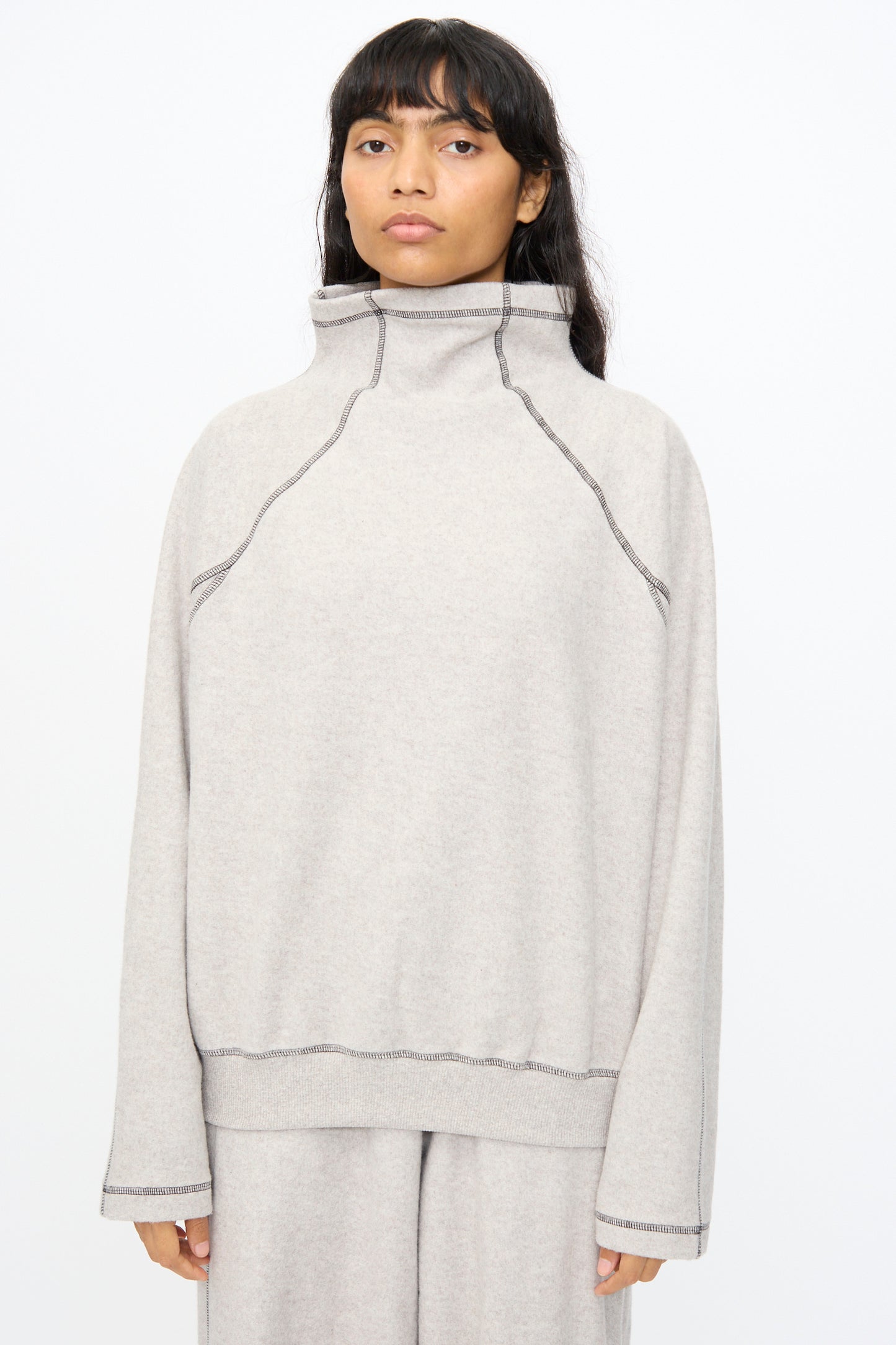 Someone wearing the Baserange Fold Turtleneck in Grey Melange—an oversized organic cotton sweater with exposed seams—stands against a plain white background.