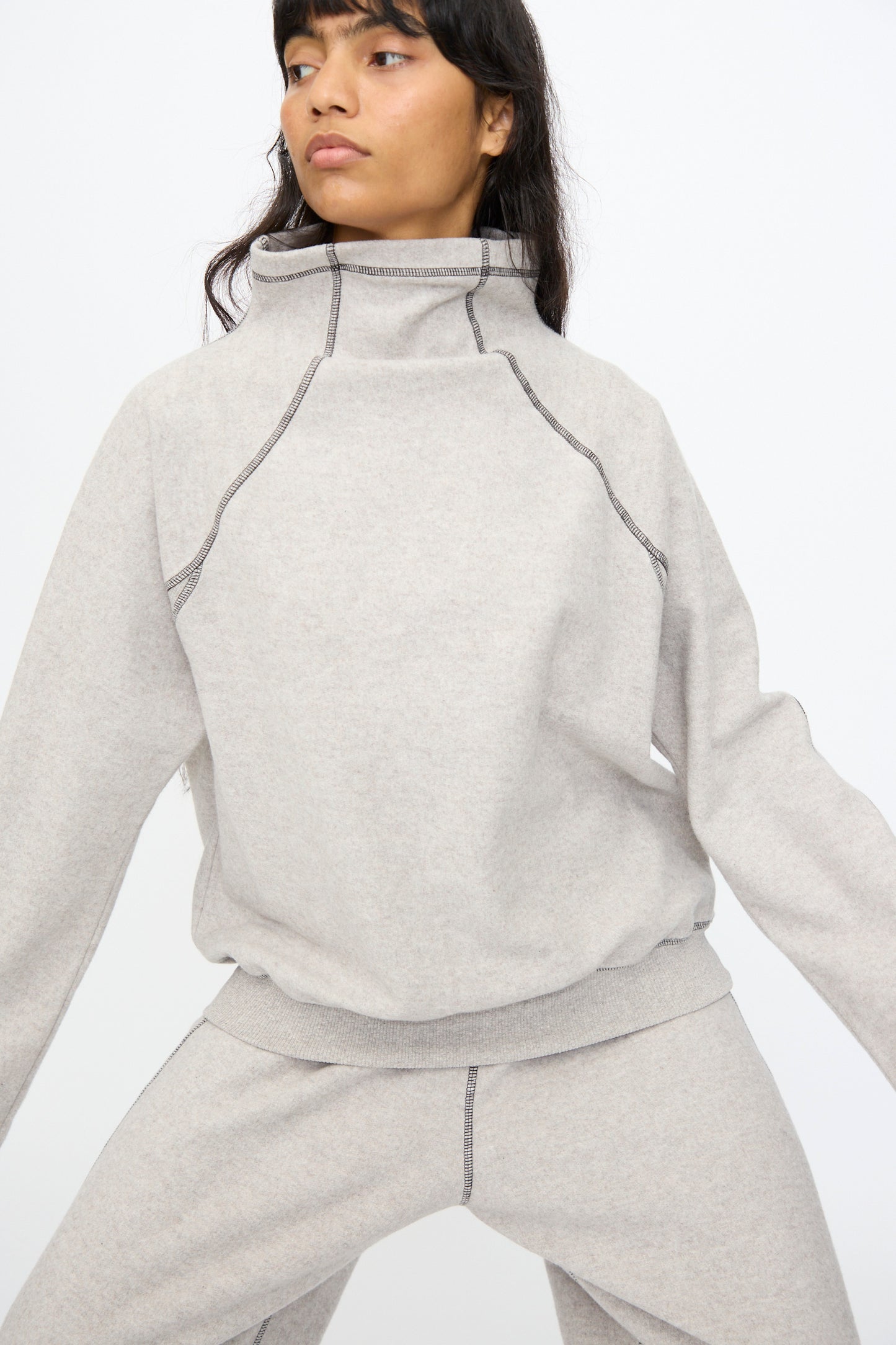 A person poses against a plain white background wearing the Baserange Fold Turtleneck in Grey Melange, a light gray organic cotton sweatshirt with exposed seam details.