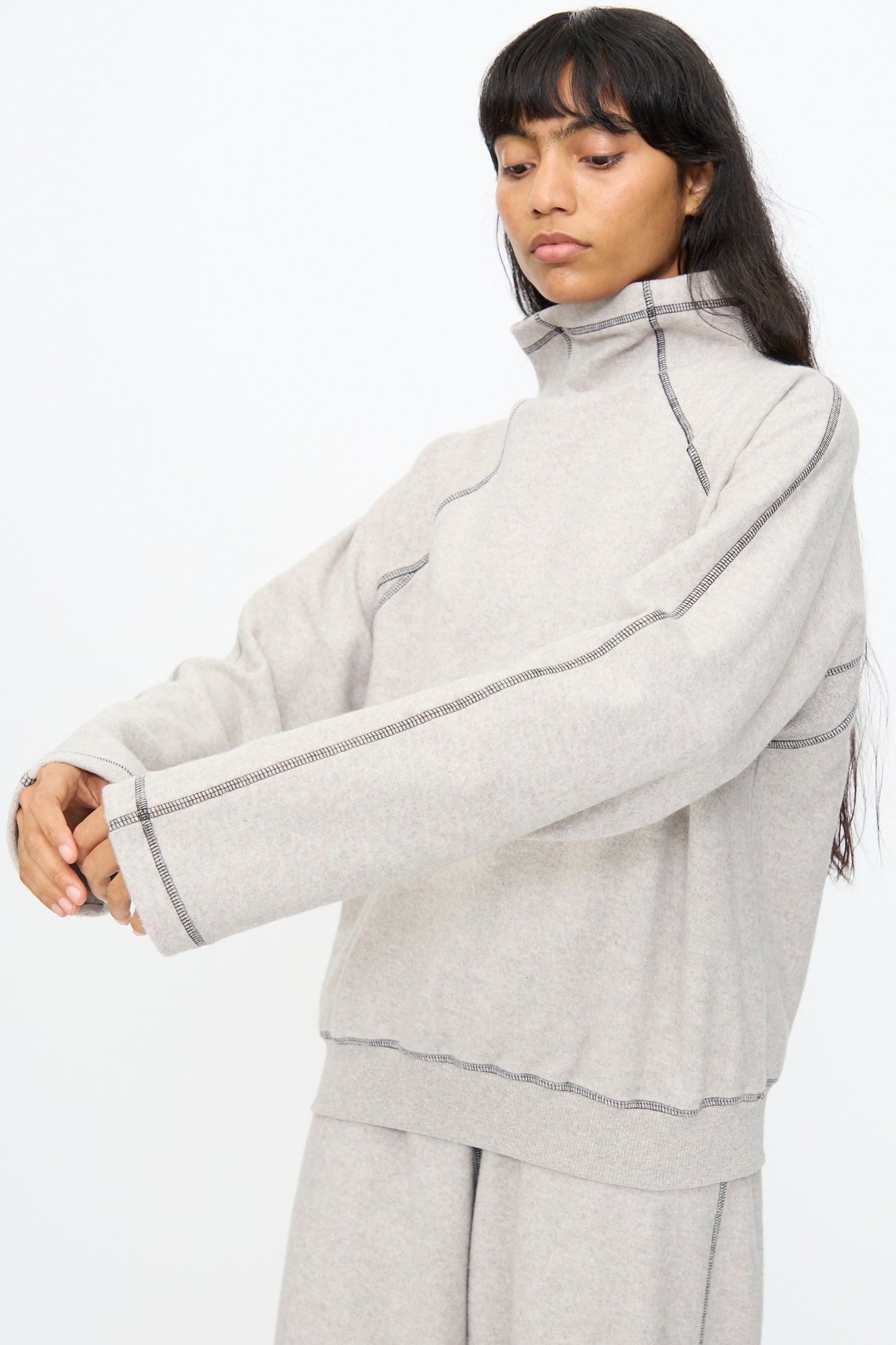 A person wears the Baserange Fold Turtleneck in Grey Melange, an oversized recycled cotton sweater with exposed seams, paired with light gray pants, standing against a plain white background.