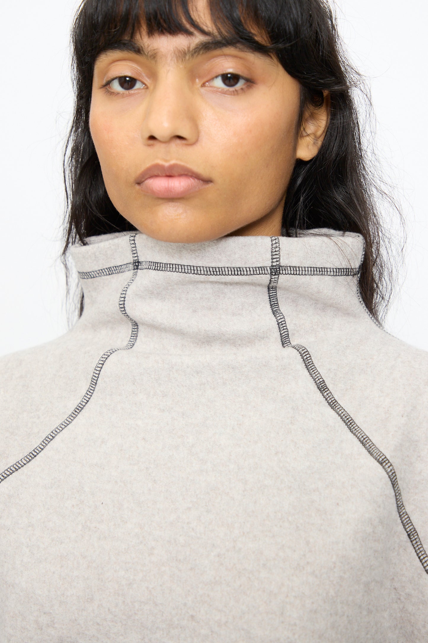 A person with long dark hair wears the Baserange Fold Turtleneck in Grey Melange—an organic cotton top with visible black stitching—while looking directly at the camera.