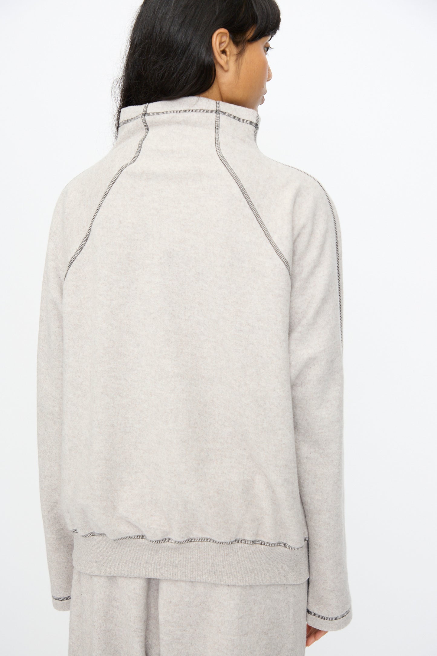 A person with long dark hair is shown from the back wearing the Baserange Fold Turtleneck in Grey Melange, featuring visible seams and matching pants, set against a plain white background.