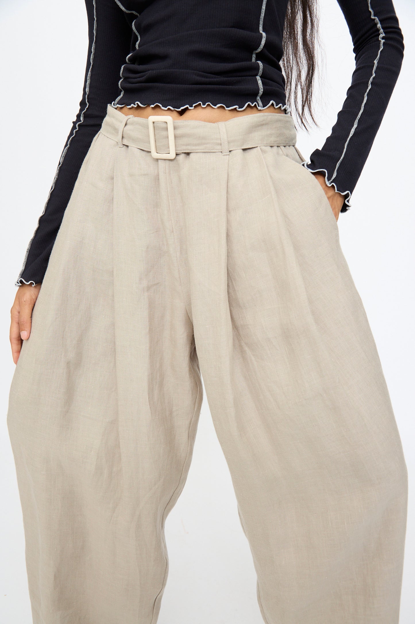 Someone is wearing Baserange's Frame Pants in Concrete—high-waisted, wide-leg beige pants with a belt—paired with a long-sleeve black top featuring white stitching.