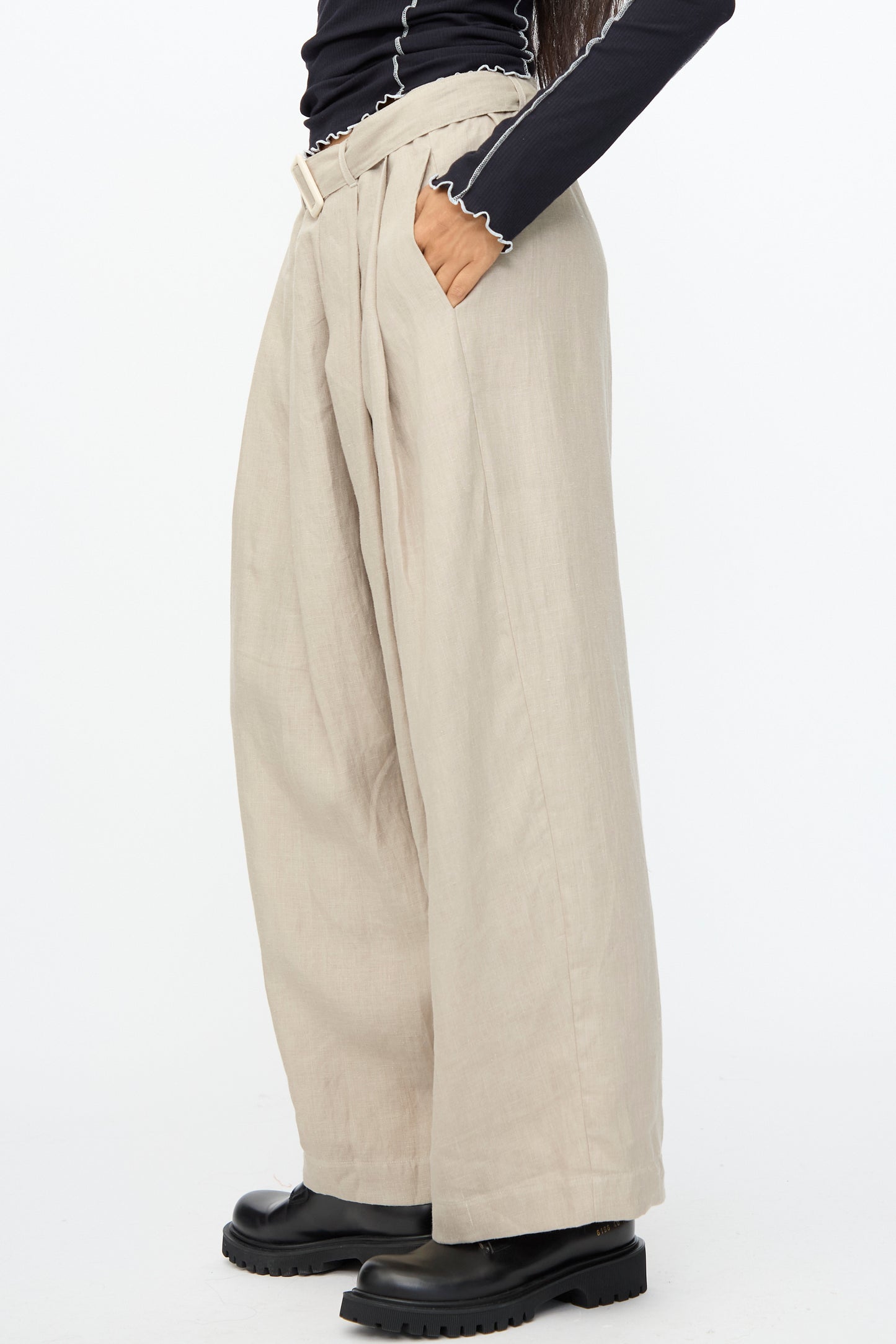Person wearing Baserange Frame Pants in Concrete with a fabric belt, paired with a black long-sleeve ruffled top and black chunky boots, standing against a white background.