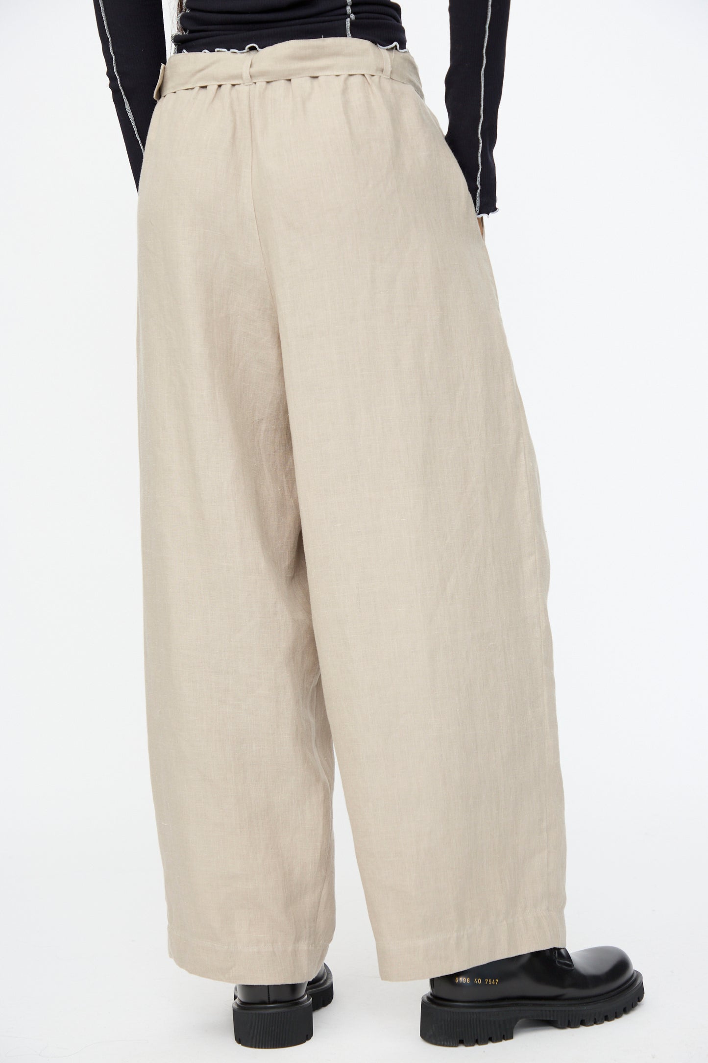 Model wears Baserange Frame Pants in Concrete—loose, wide-leg trousers—paired with black boots, seen from the back against a plain white background.