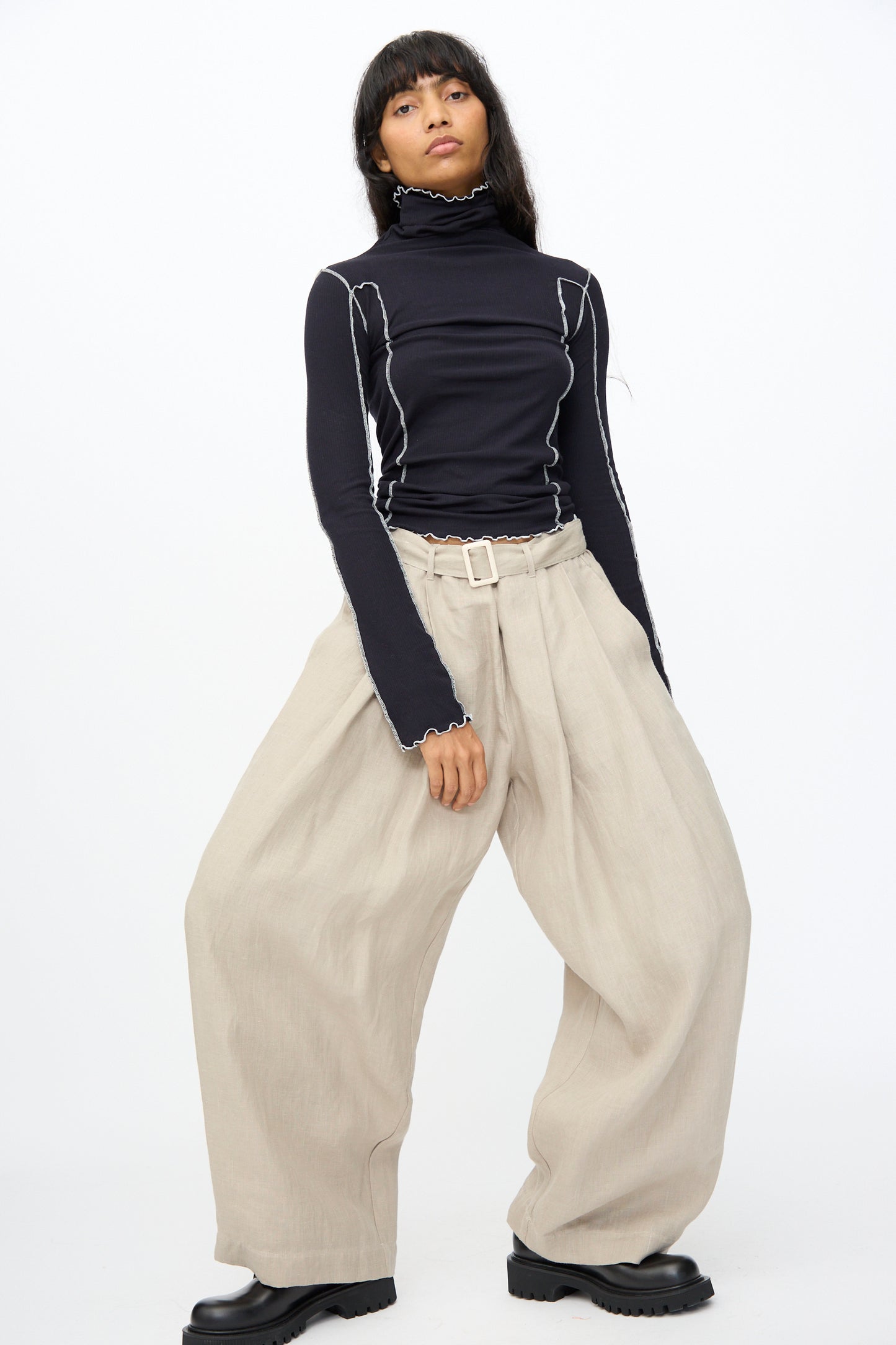 Model wears Baserange Frame Pants in Concrete, featuring a high waist and wide legs, paired with a black turtleneck with white stitching, standing against a plain white background.