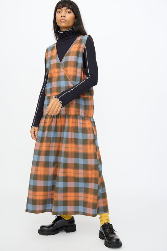 A person wears the Baserange Garlin Dress in Frame Plaid over a black turtleneck, paired with yellow socks and black shoes, standing against a plain white background.