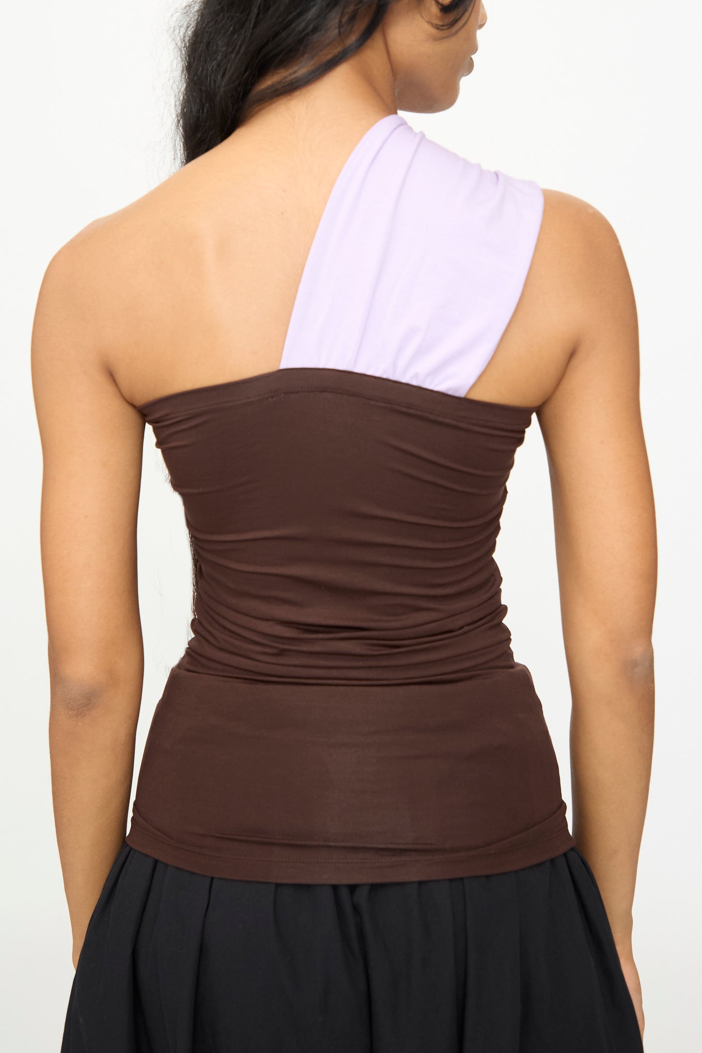 A person seen from the back wearing the Baserange Geo Tank in Tactile One Purple, a brown, asymmetrical one-shoulder top featuring a light purple single shoulder strap.