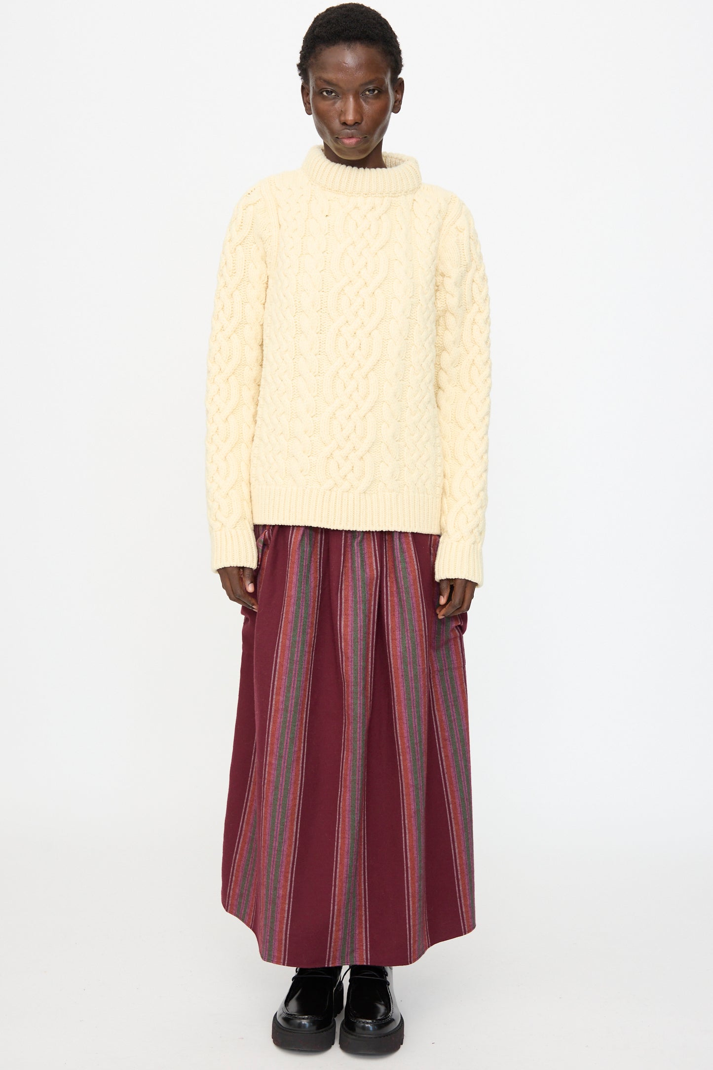 A person stands against a white background, wearing the Baserange Lens Sweater in Chaume Sand, paired with a long maroon skirt featuring vertical stripes and black platform shoes.