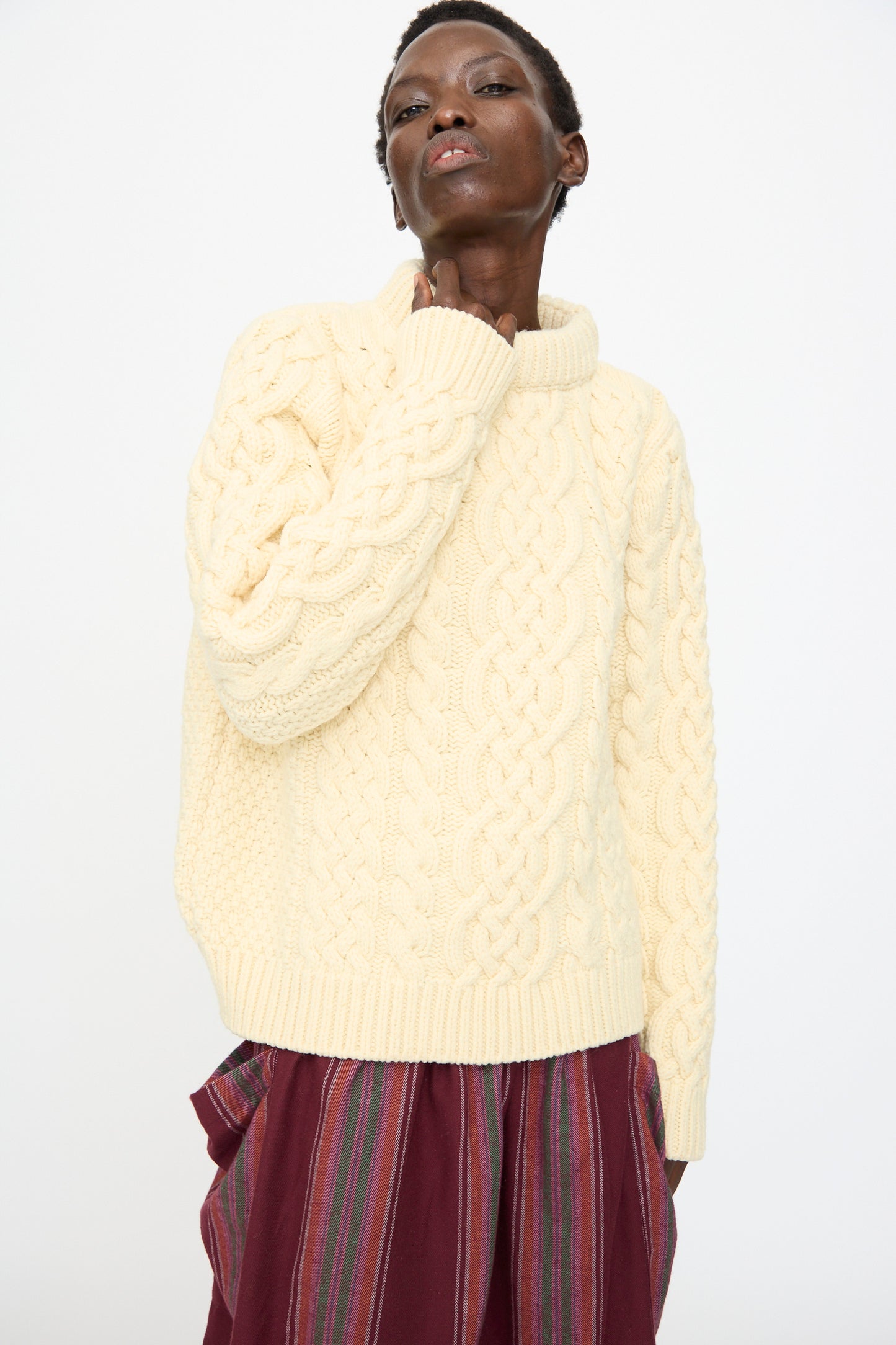 Person wearing the Baserange Lens Sweater in Chaume Sand, paired with a maroon striped skirt, standing against a plain white background.