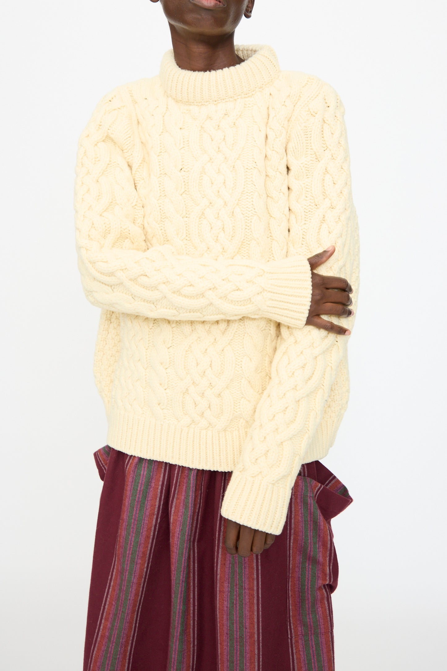 A person wears the Baserange Lens Sweater in Chaume Sand paired with a maroon skirt featuring vertical stripes, set against a plain background.