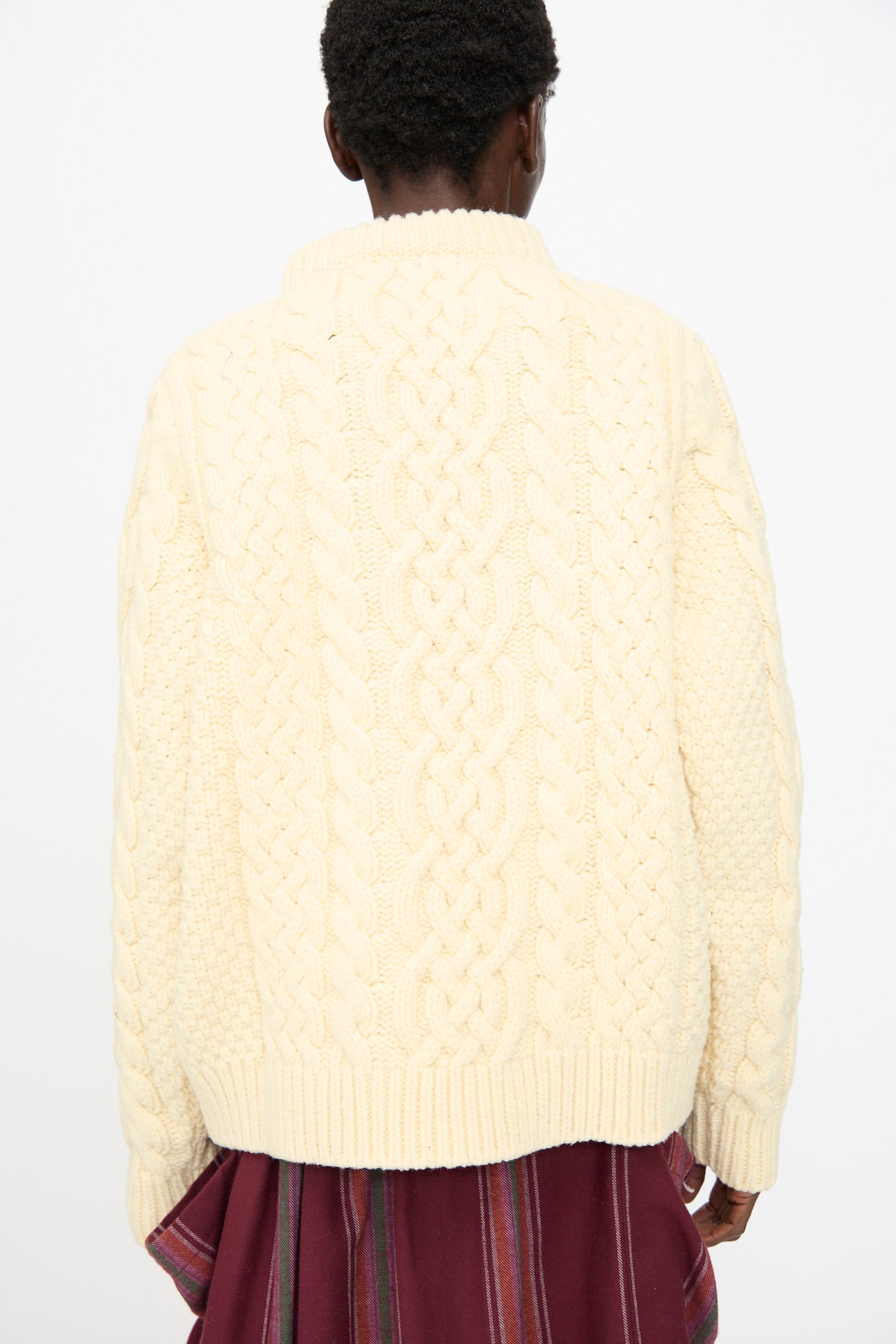 A person faces away, wearing the Baserange Lens Sweater in Chaume Sand, a cream-colored chunky knit merino wool top, paired with a maroon vertically striped skirt, set against a plain white background.