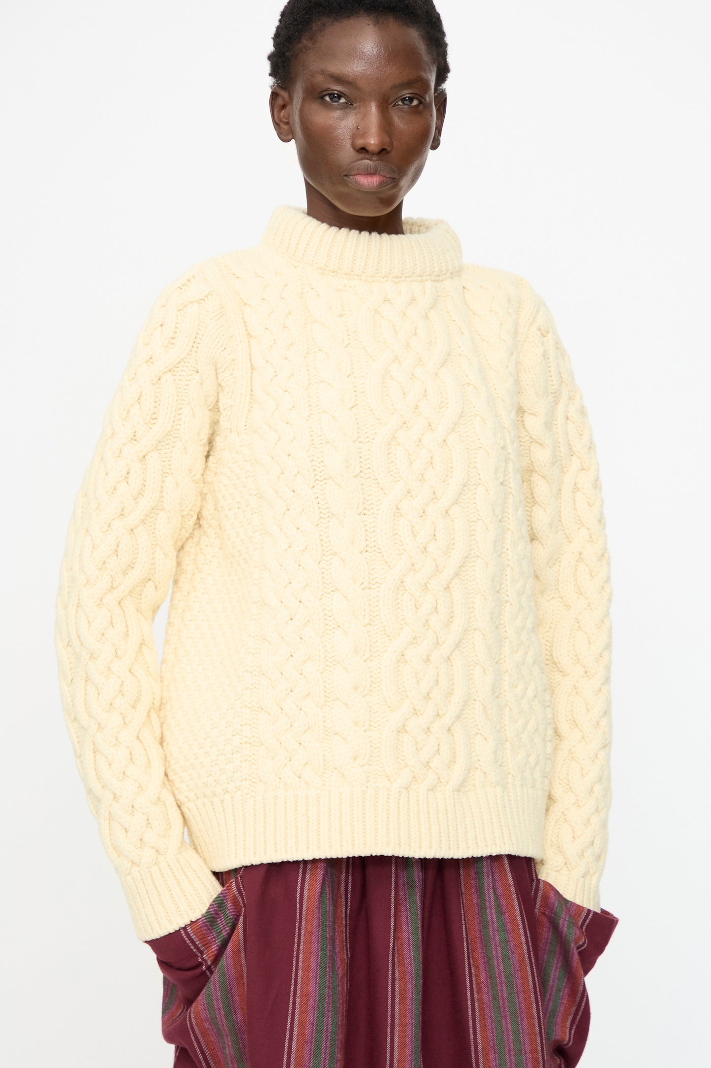 A person wearing the Baserange Lens Sweater in Chaume Sand and a maroon vertically striped skirt, standing against a plain white background.