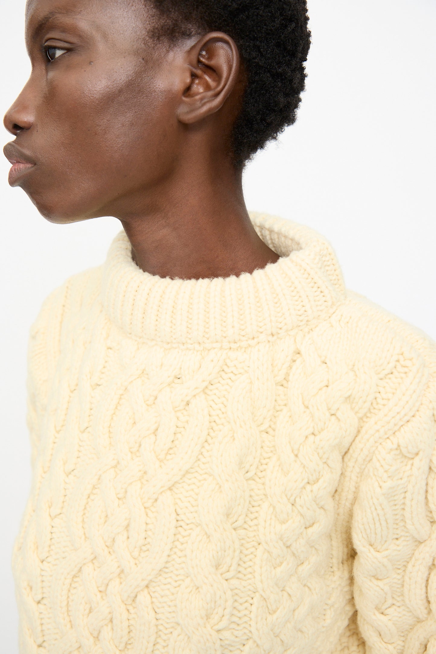 A person in profile wears the Baserange Lens Sweater in Chaume Sand, a chunky knit merino wool turtleneck, set against a plain light background.