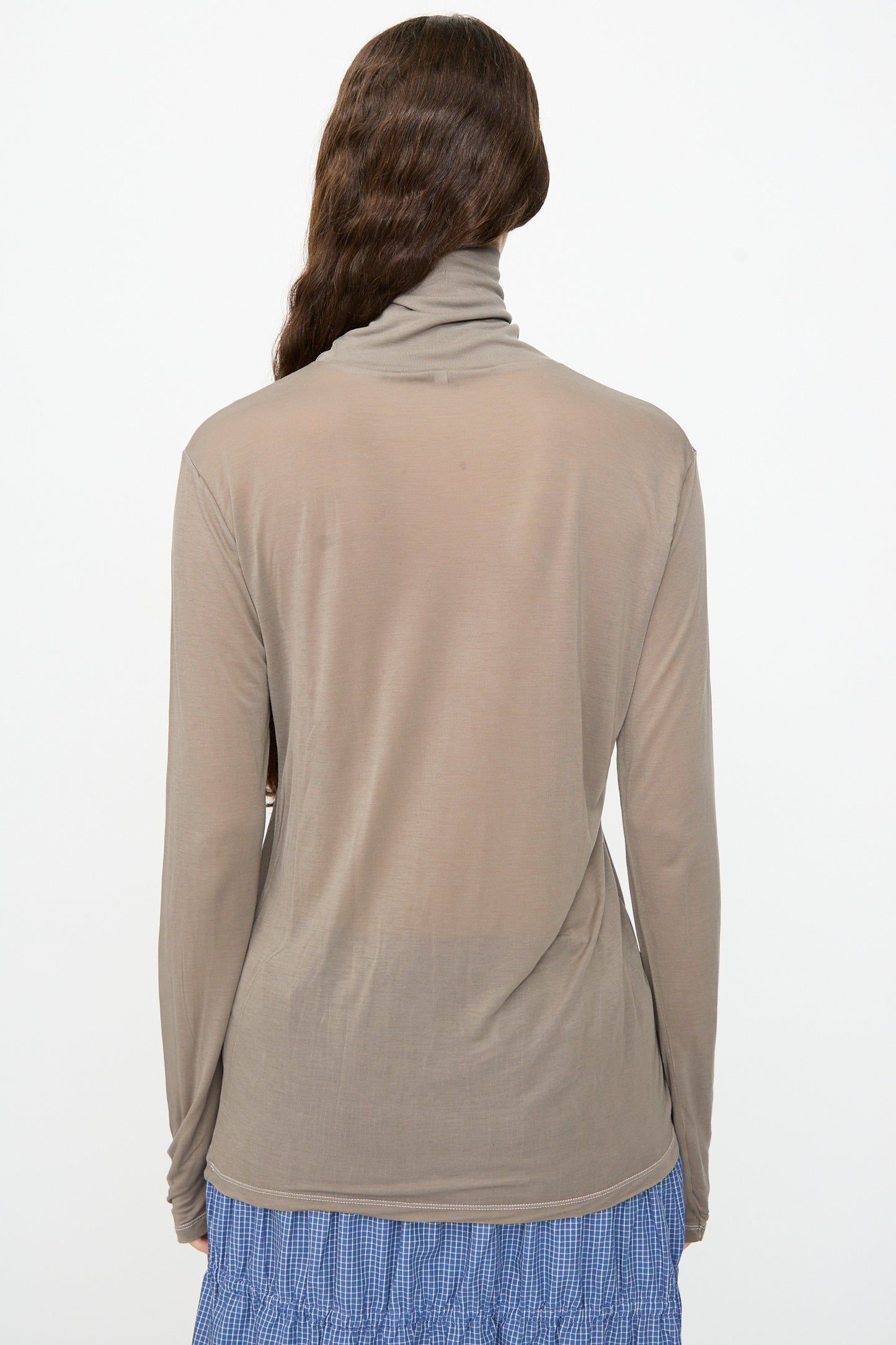 A person with long wavy brown hair is shown from the back against a plain white background, wearing the Baserange Long Sleeve Turtleneck in Gilda Grey paired with a blue plaid skirt.