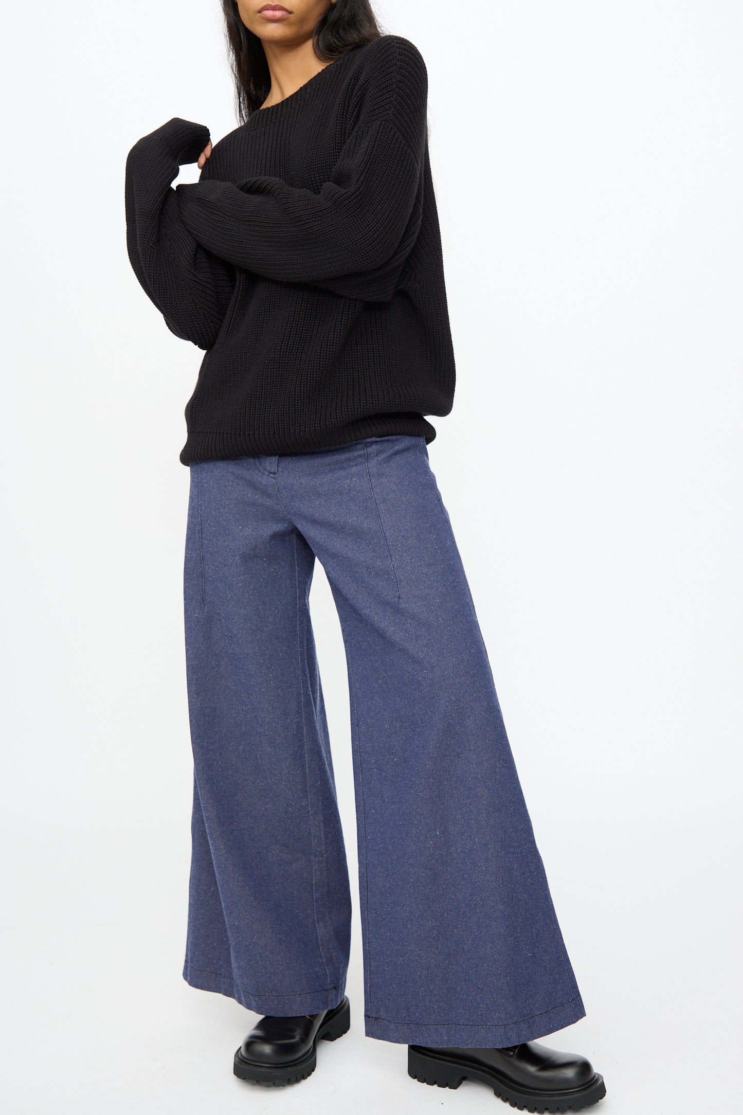 A person wearing the Baserange Mea Knit Pullover in Black, styled with wide-leg blue jeans and black platform shoes, stands against a plain white background.