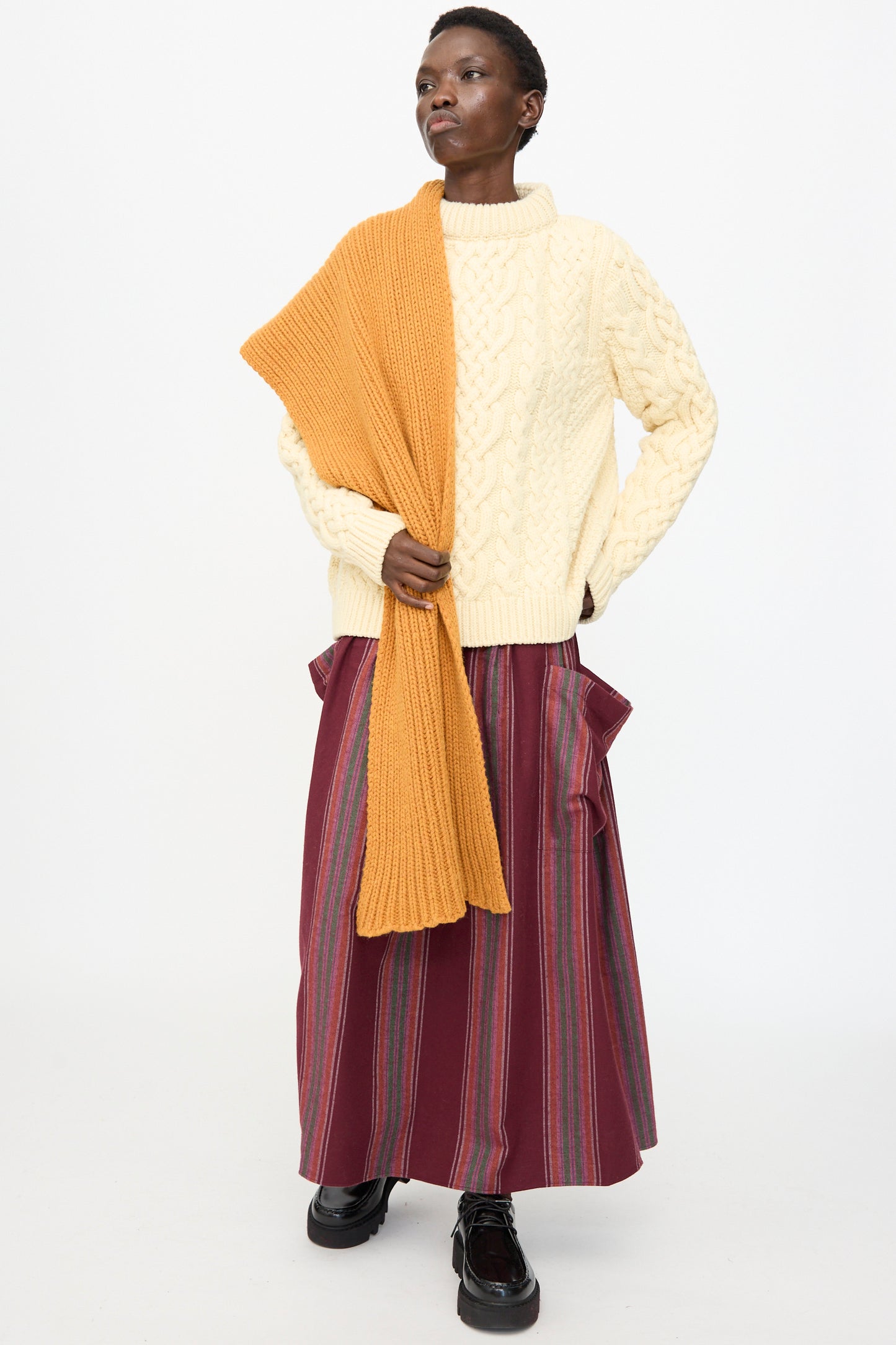 A person stands against a plain white background wearing a cream cable-knit sweater, long striped maroon skirt, black shoes, and the Baserange Mea Scarf in Clair Orange made from recycled wool.