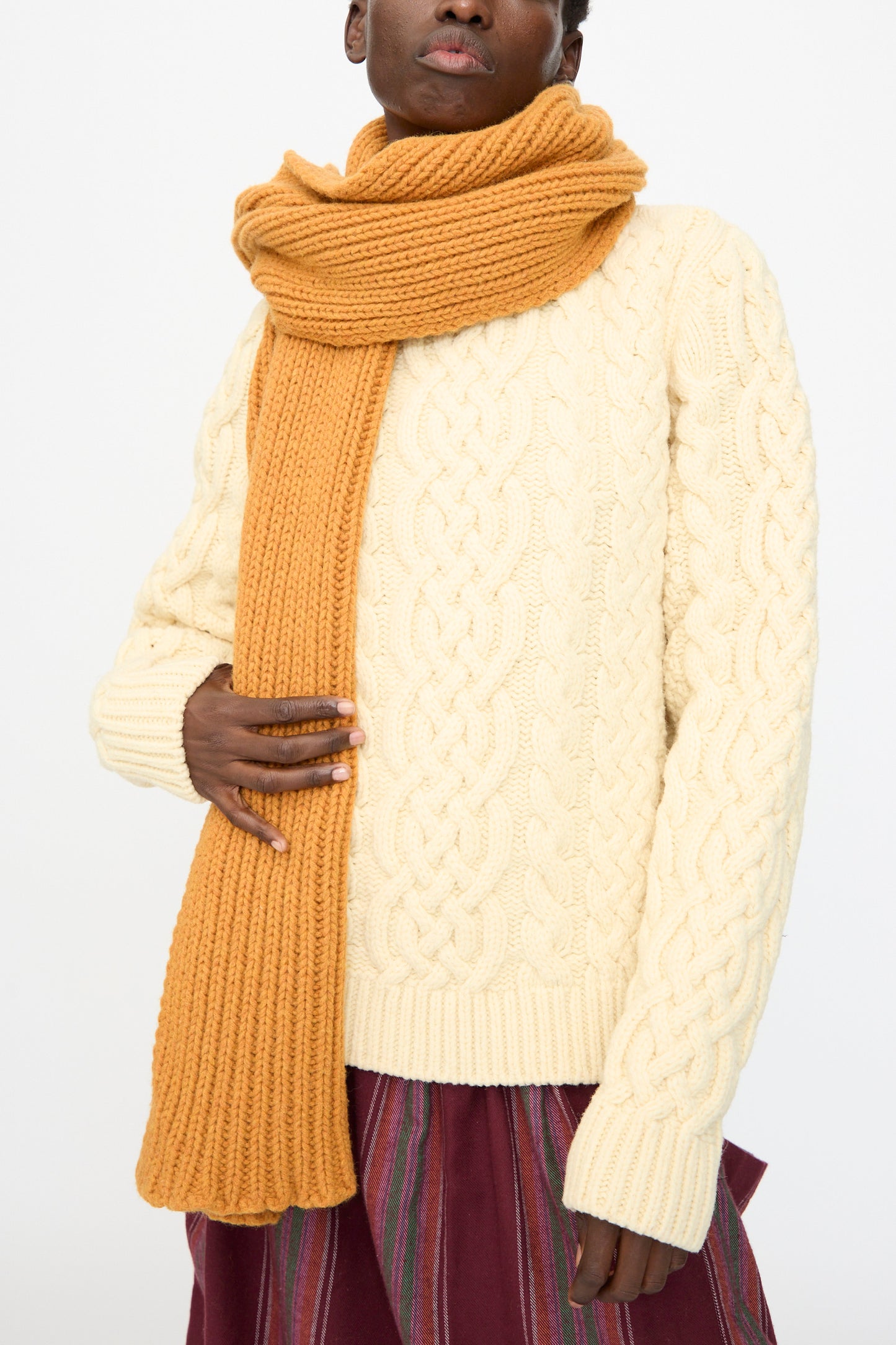 A person wearing a cream cable-knit sweater, Baserange's Mea Scarf in Clair Orange, and a vertically striped skirt stands against a plain white background.