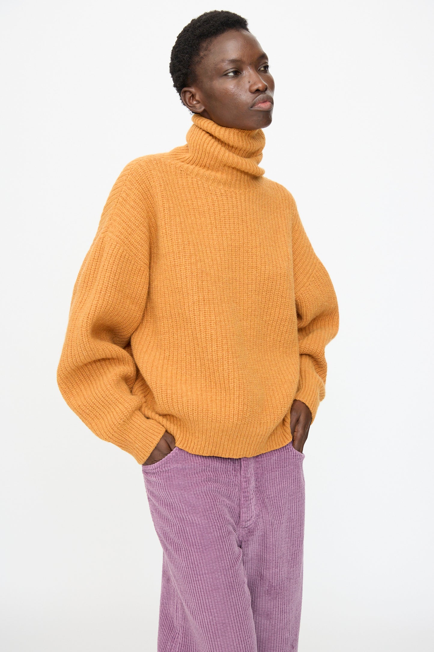 A person wears the Baserange Mea Turtleneck in Clair Orange with light purple corduroy pants, hands in pockets, against a plain white background.