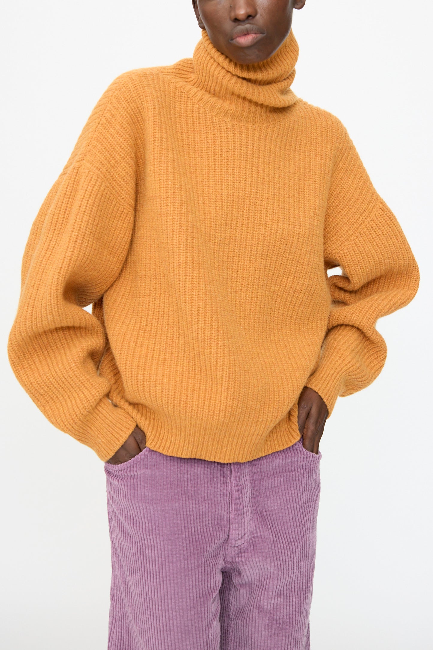 A person wears the Baserange Mea Turtleneck in Clair Orange with light purple corduroy pants, standing with hands in pockets against a plain background.