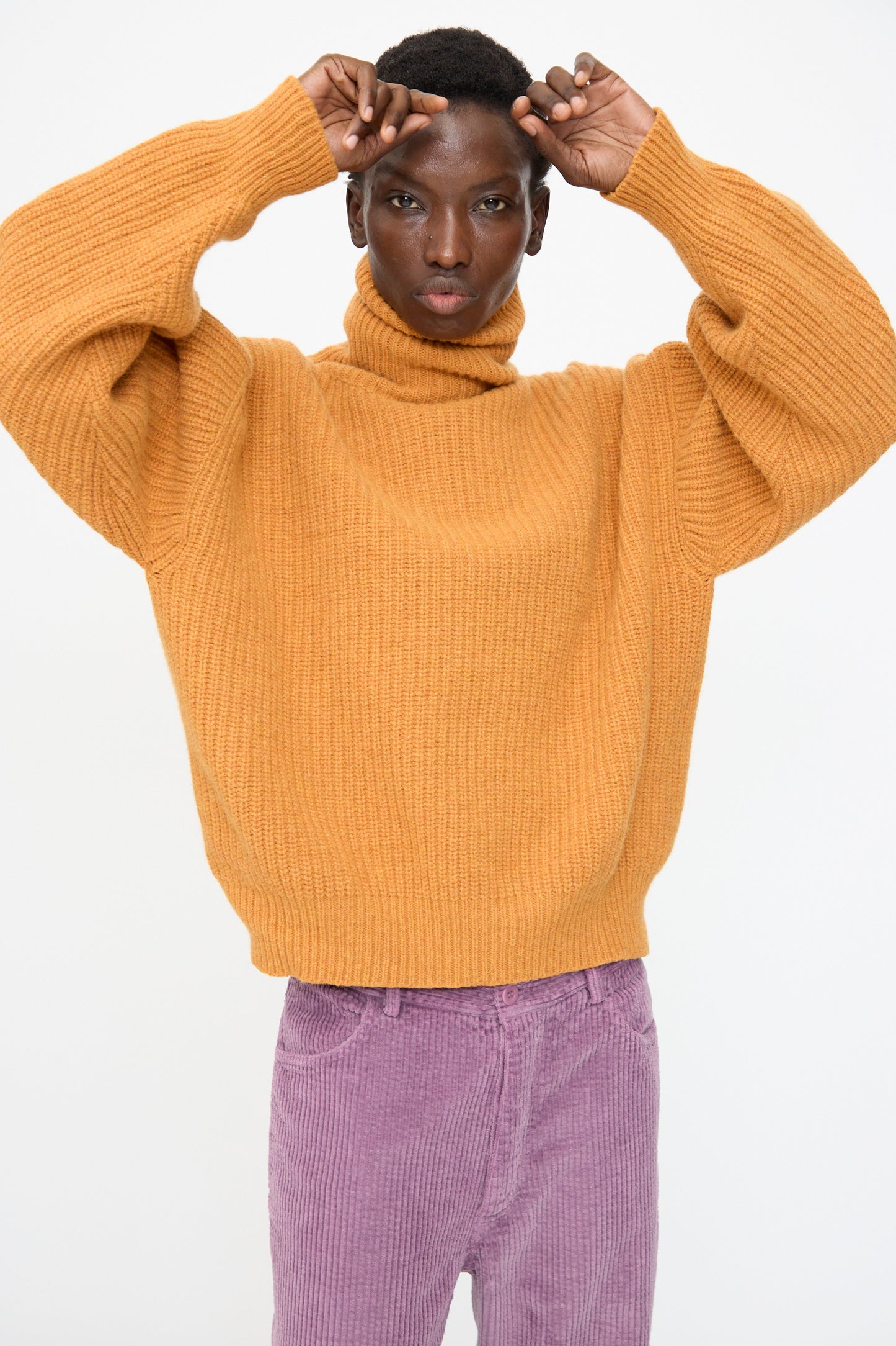 A person wearing the Baserange Mea Turtleneck in Clair Orange poses with hands raised near their head, paired with lavender corduroy pants, against a plain light background.