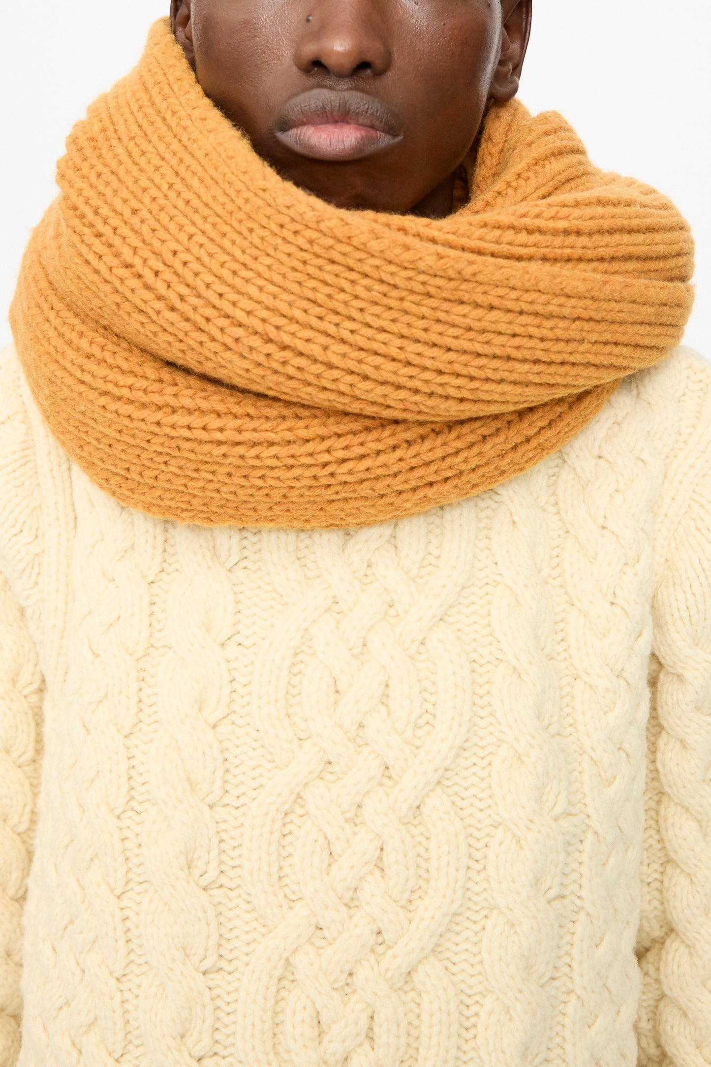 A person is shown from shoulders to mouth wearing the Baserange Mea Scarf in Clair Orange, a chunky knit made from recycled wool, paired with a cream cable knit sweater.