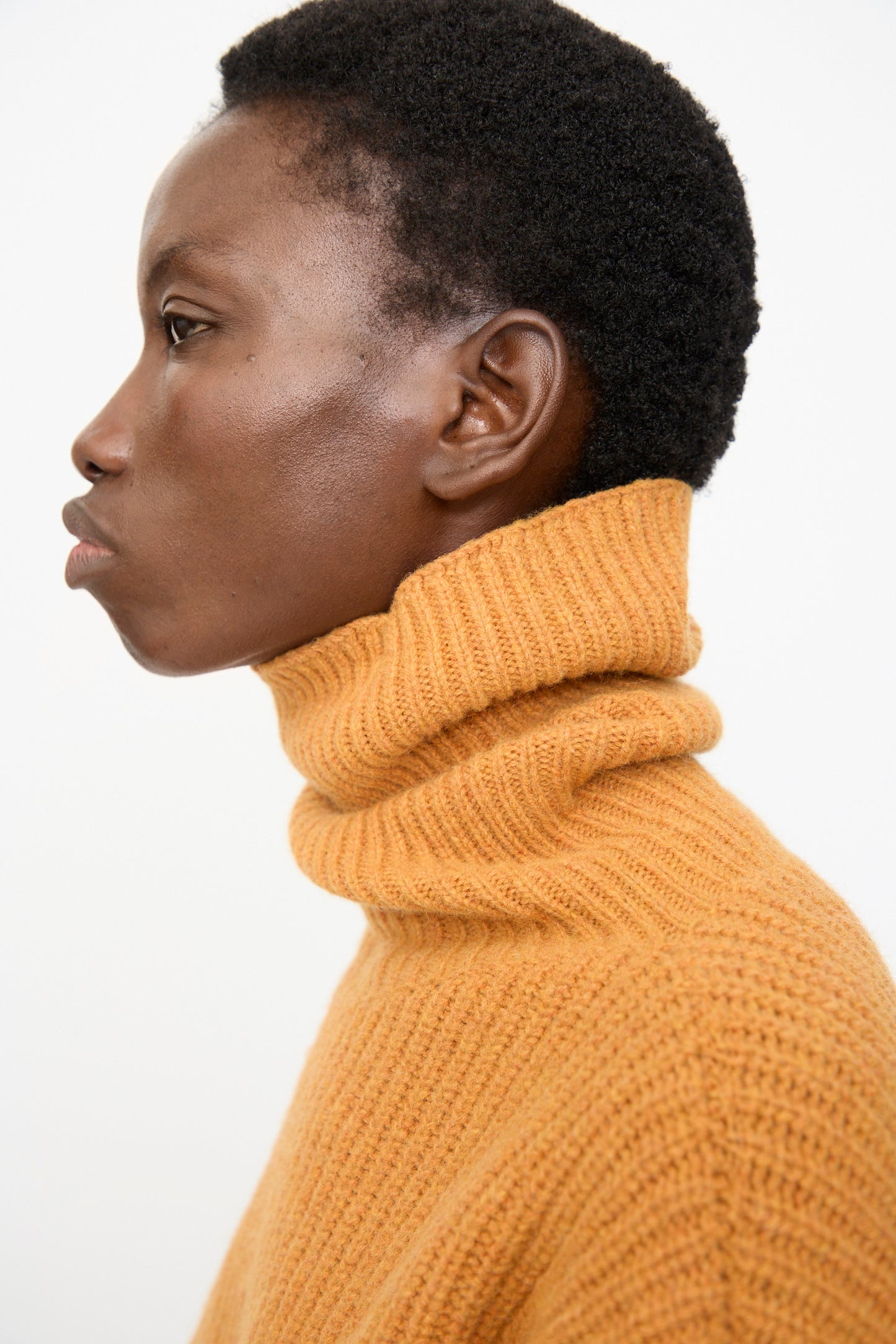A person with short curly hair wears the Baserange Mea Turtleneck in Clair Orange, shown in profile against a plain white background.
