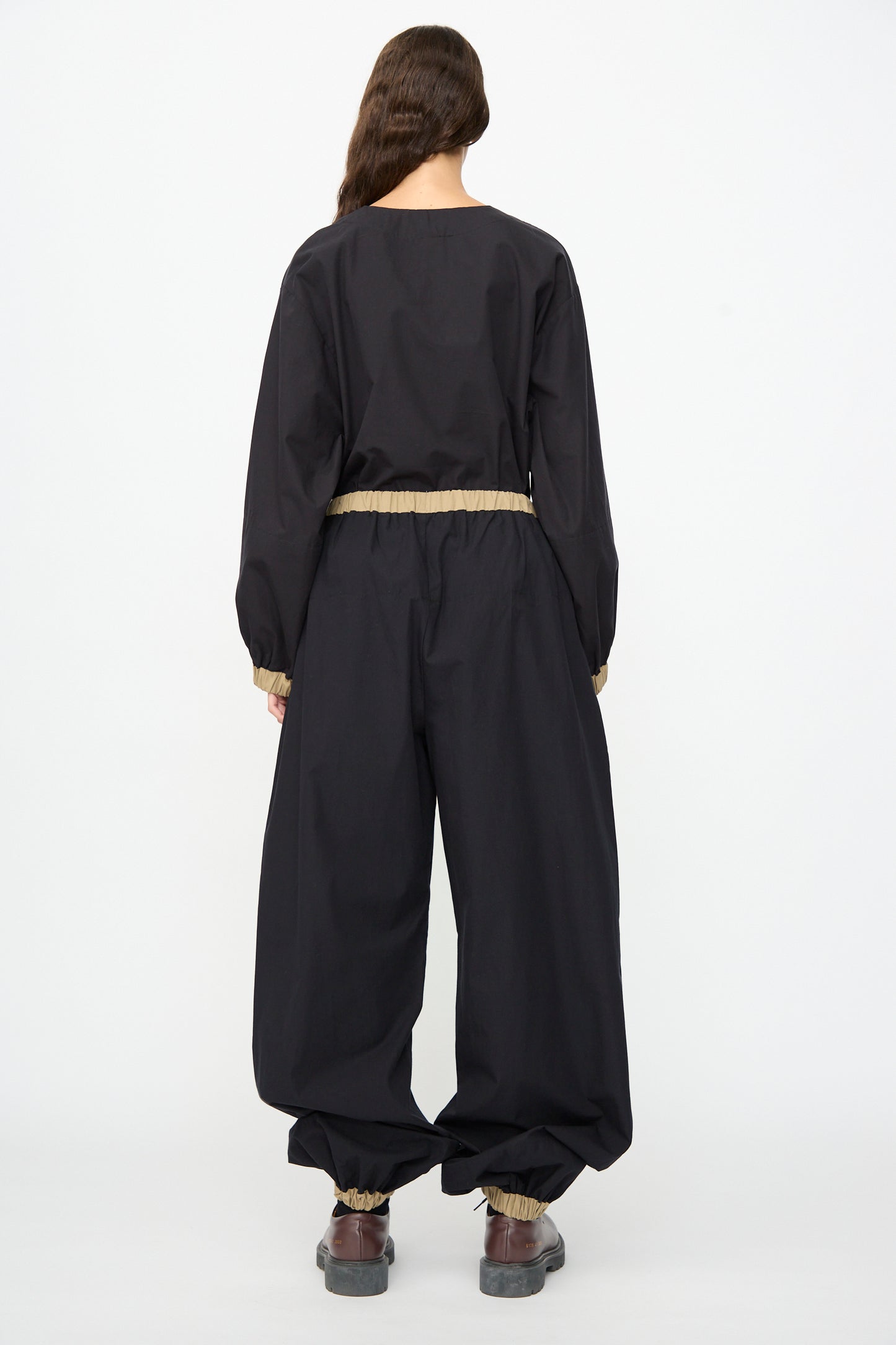 A person with long brown hair stands with their back to the camera, wearing the Baserange Moy Pant in Black Khaki—loose-fitting black cotton poplin pants featuring an elastic waistband and cuffs—paired with brown shoes.