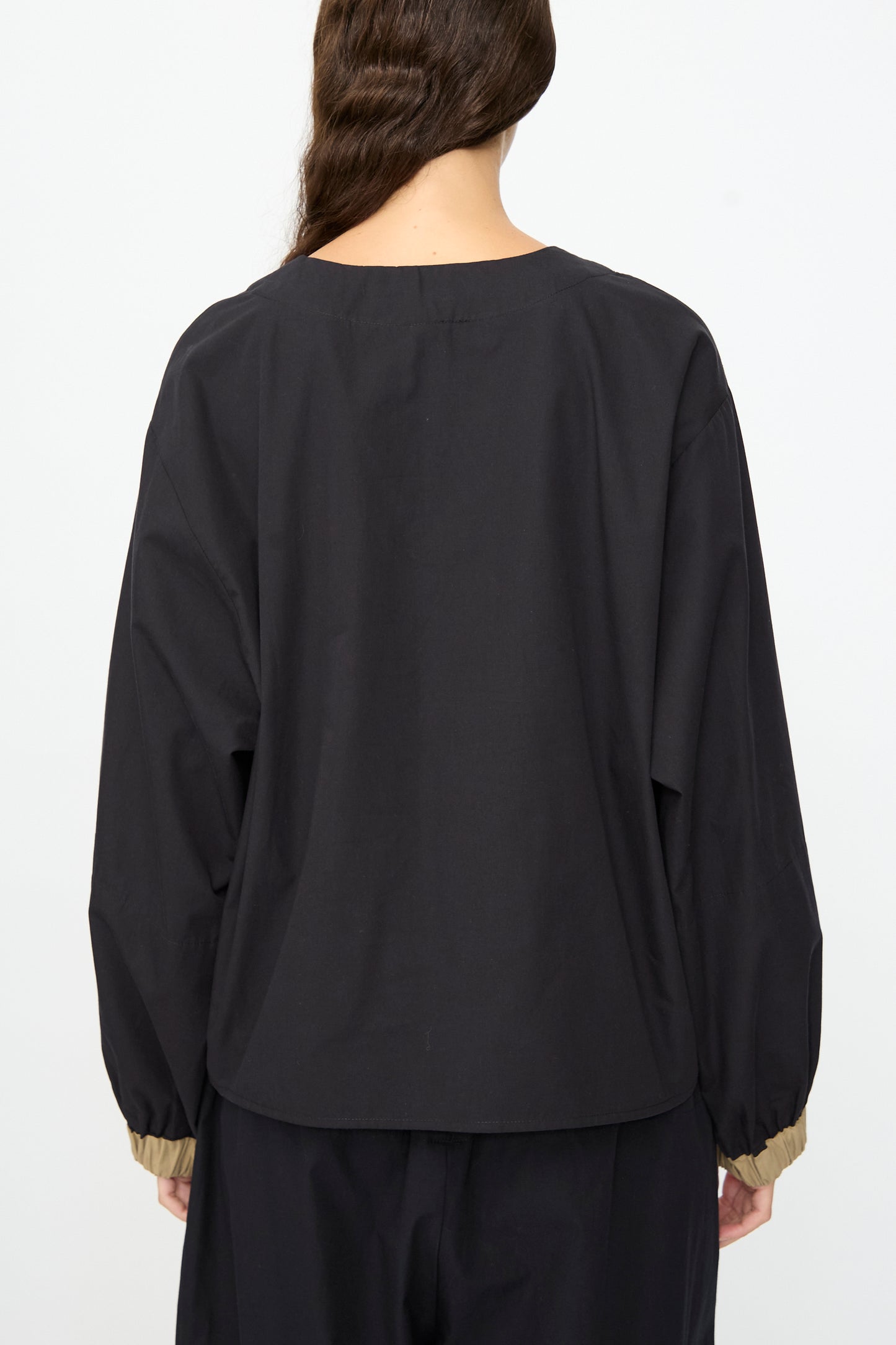 A person with long brown hair is shown from the back, wearing the Baserange Moy Top in Black Khaki—a relaxed fit black long-sleeve top with beige cuffs—paired with matching black pants.